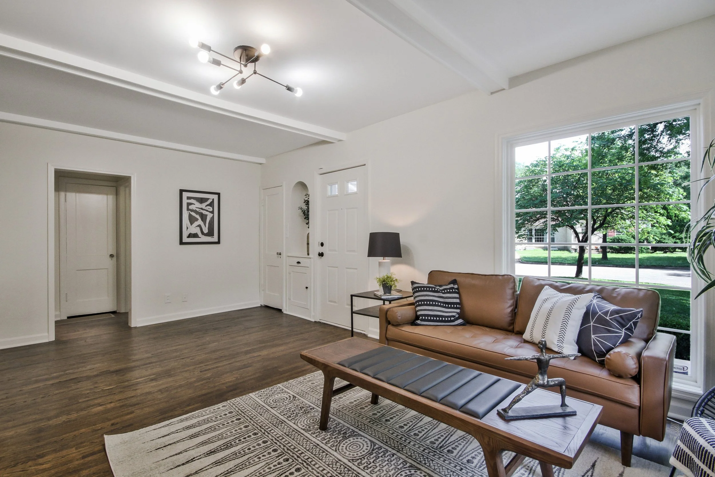 Living room with a brown leather sofa, black and white pillows, a wooden bench, a black side table with a lamp and small plant, large window with green trees outside, wooden flooring, white walls, and ceiling light fixture.