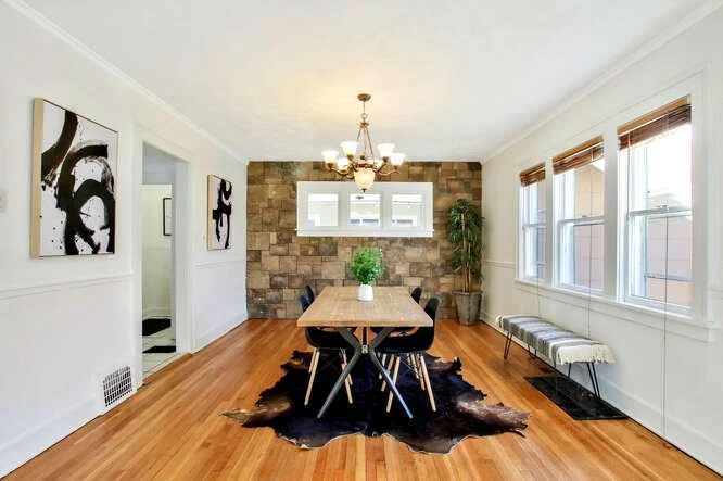 Dining room with a wooden table, four black chairs, and a black cowhide rug, with a stone accent wall, chandelier, window, and a bench.
