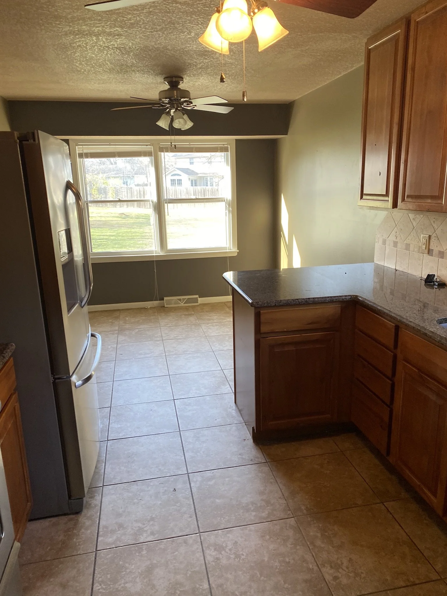 A kitchen with tan tiled floor, wooden cabinets, a granite countertop, a window letting in natural light, a ceiling fan with lights, and a ceiling light fixture.