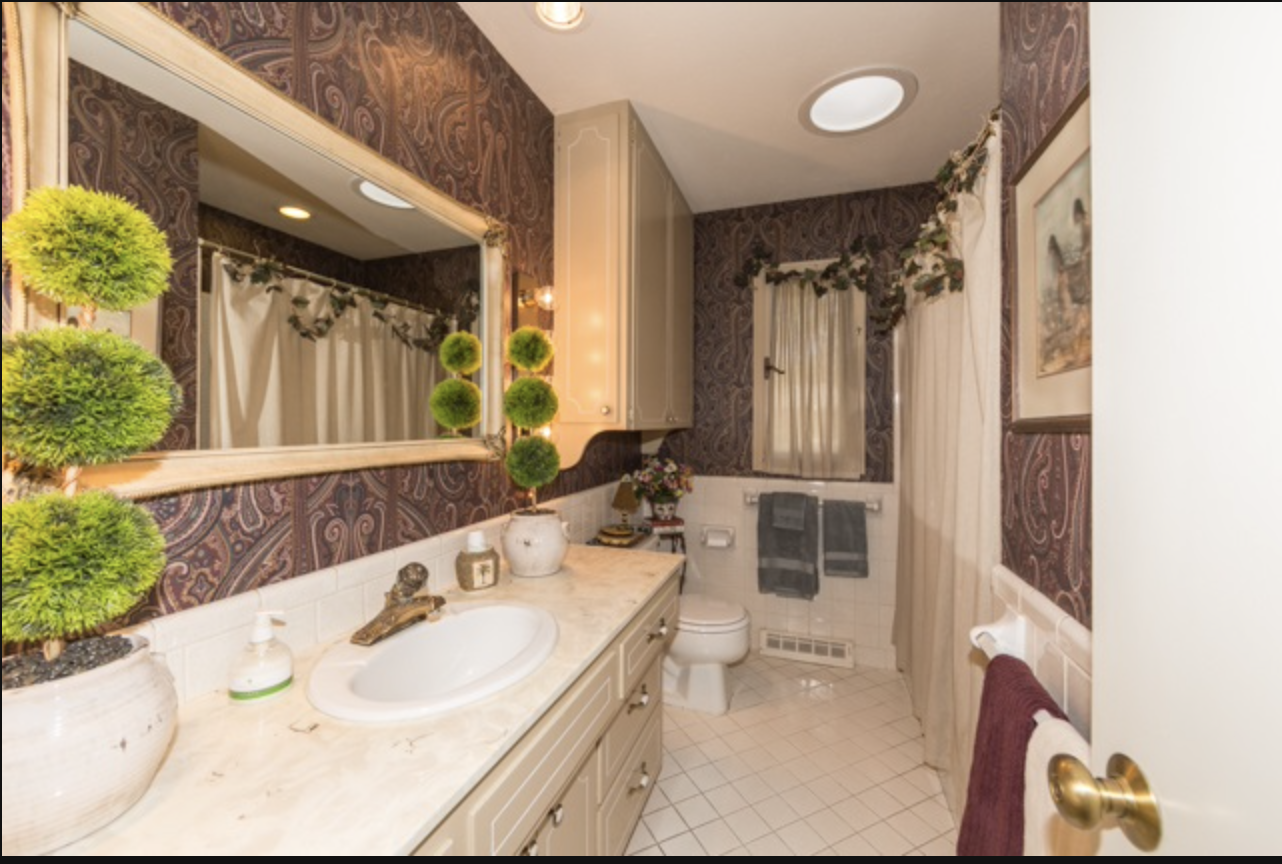 Bathroom with paisley wallpaper, beige cabinetry, large mirror, white countertop, potted topiary plants, shower with curtain, window, towels, framed artwork, and gold doorknob.