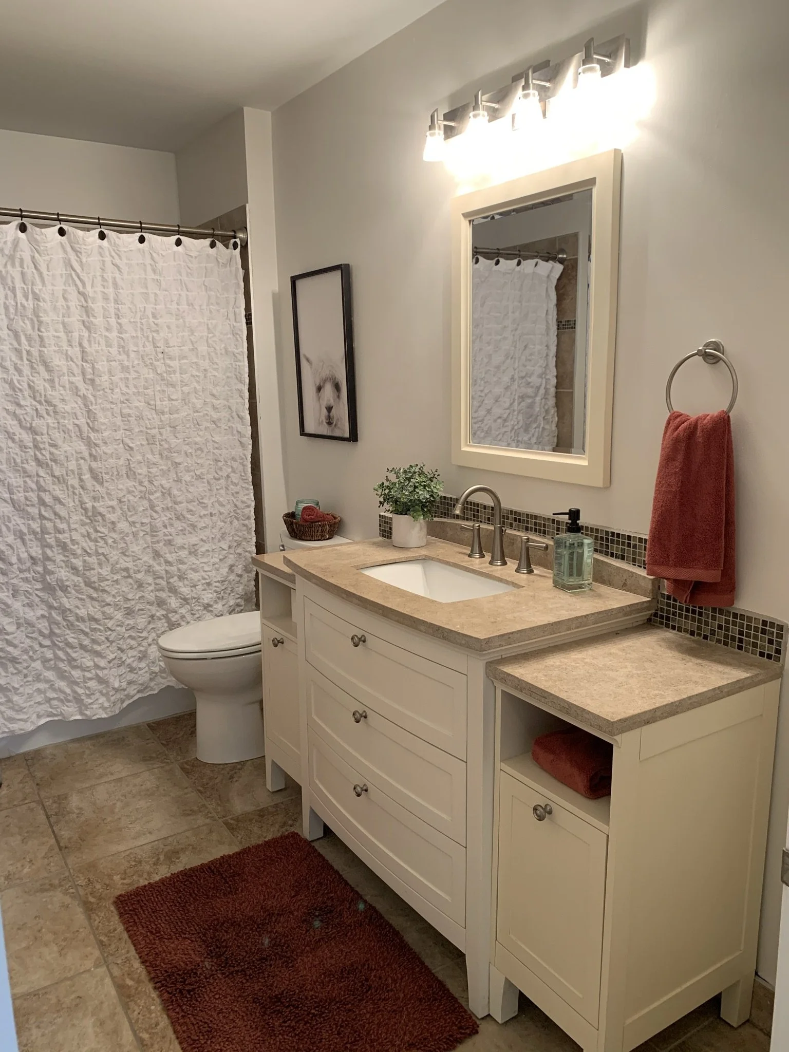 Bathroom with white vanity, beige countertop, mirror, and a towel ring with a red towel. Shower with white curtain, framed picture of a llama, and bathroom essentials on the counter.