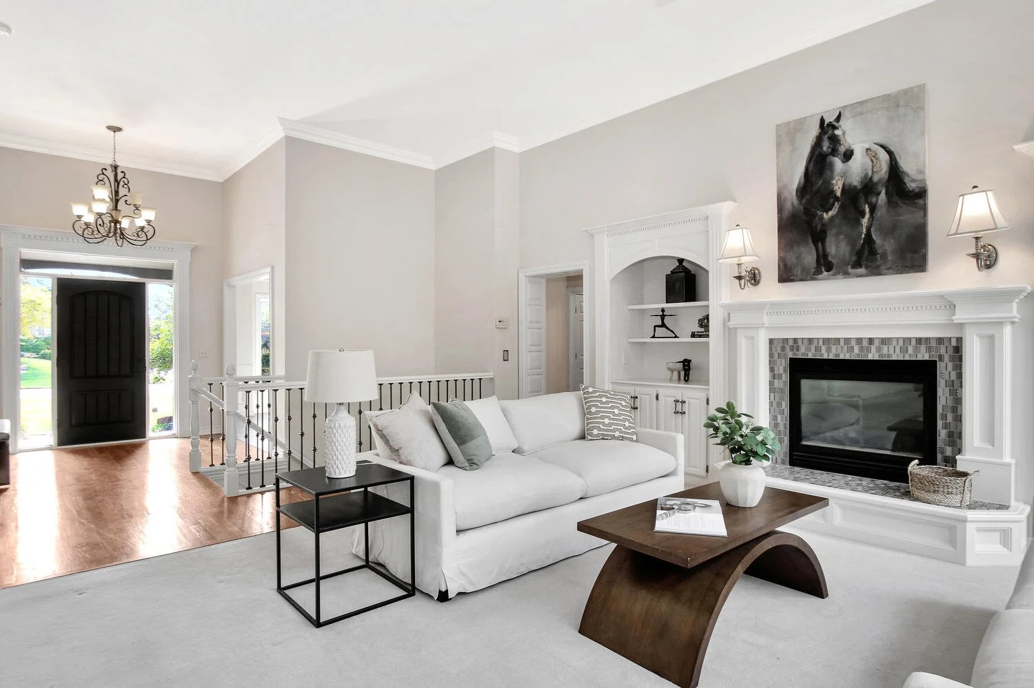 Living room with white sofa, wooden coffee table, fireplace with a painting of a horse, built-in shelves, and wall sconces in a neutral color scheme.