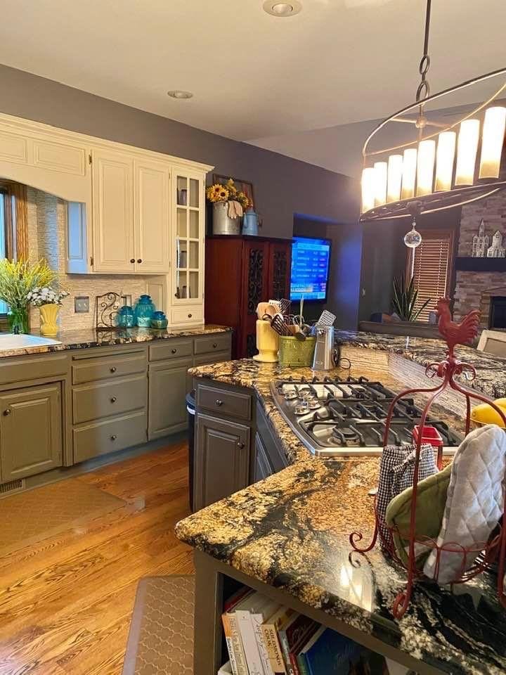 A kitchen with granite countertops, white and gray cabinets, a small window with blinds, a vase of flowers, a TV on the wall, and a chandelier overhead. There are various kitchen utensils and decorative items on the counters, and a wooden floor.