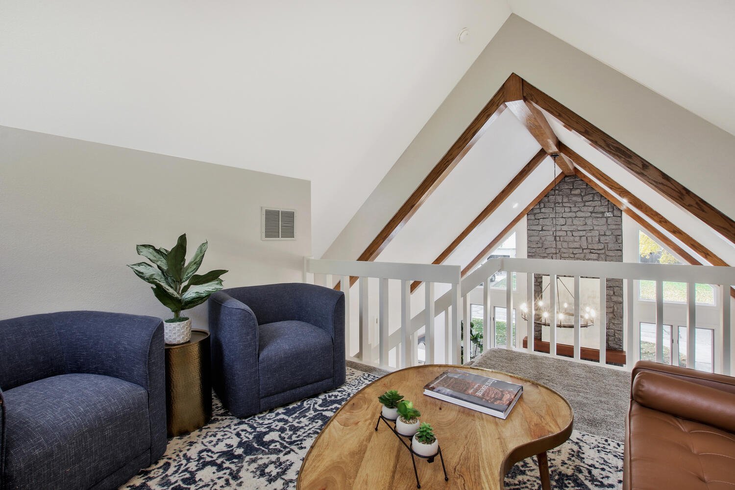 Living room with modern furniture, including a dark blue armchair, a wooden coffee table with books and small potted plants, a patterned rug, and a large window with brick wall and exposed wooden beams in the ceiling.