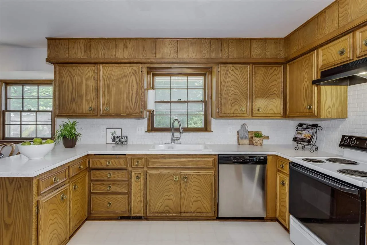 Kitchen with wooden cabinets, white countertops, a window above the sink, a potted plant, a bowl of green apples, and kitchen appliances including a dishwasher and stove.