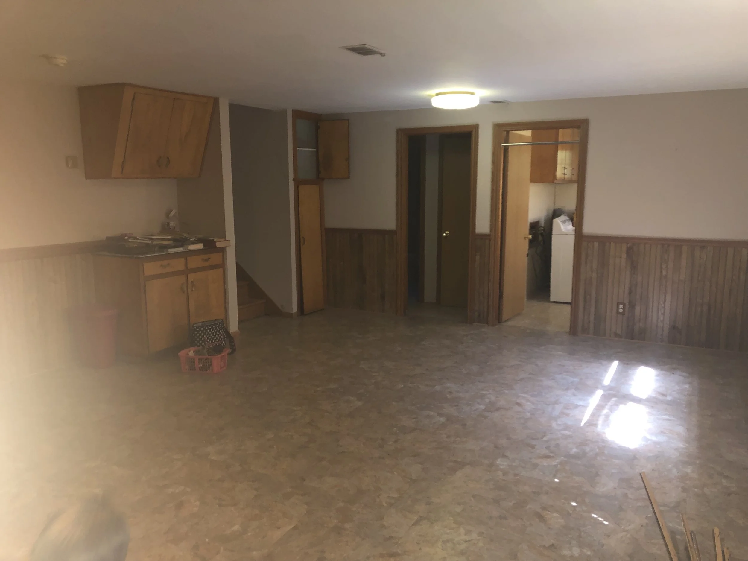 Empty room with wood paneling, open doors leading to a laundry area, and a kitchenette with cabinets, countertops, a stovetop, and a trash can.