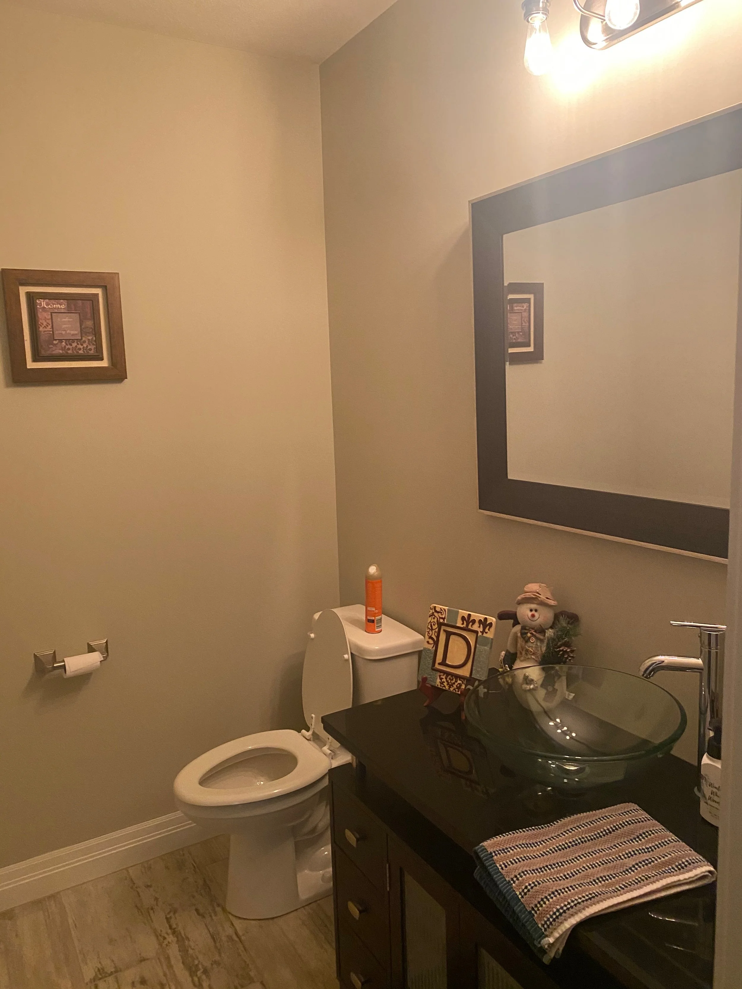 A small bathroom with a toilet, a black countertop with a glass vessel sink, a large mirror, and a wooden cabinet. Decor includes picture frames, a small snowman figurine, a hand towel, and toiletries.