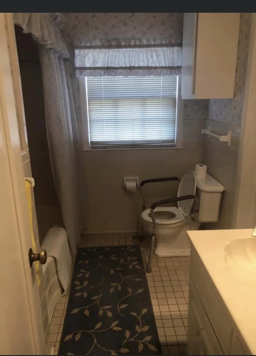 Small bathroom with floral wallpaper, window with blinds and valance, toilet with grab bars, toilet paper holder, towel rack, shower with curtain, patterned rug, and bathroom sink