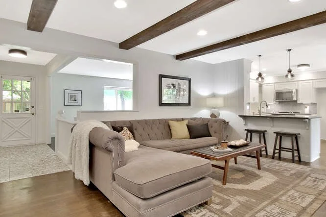 Living room with a beige sectional sofa, a wooden coffee table, and a kitchen with a breakfast bar in the background.