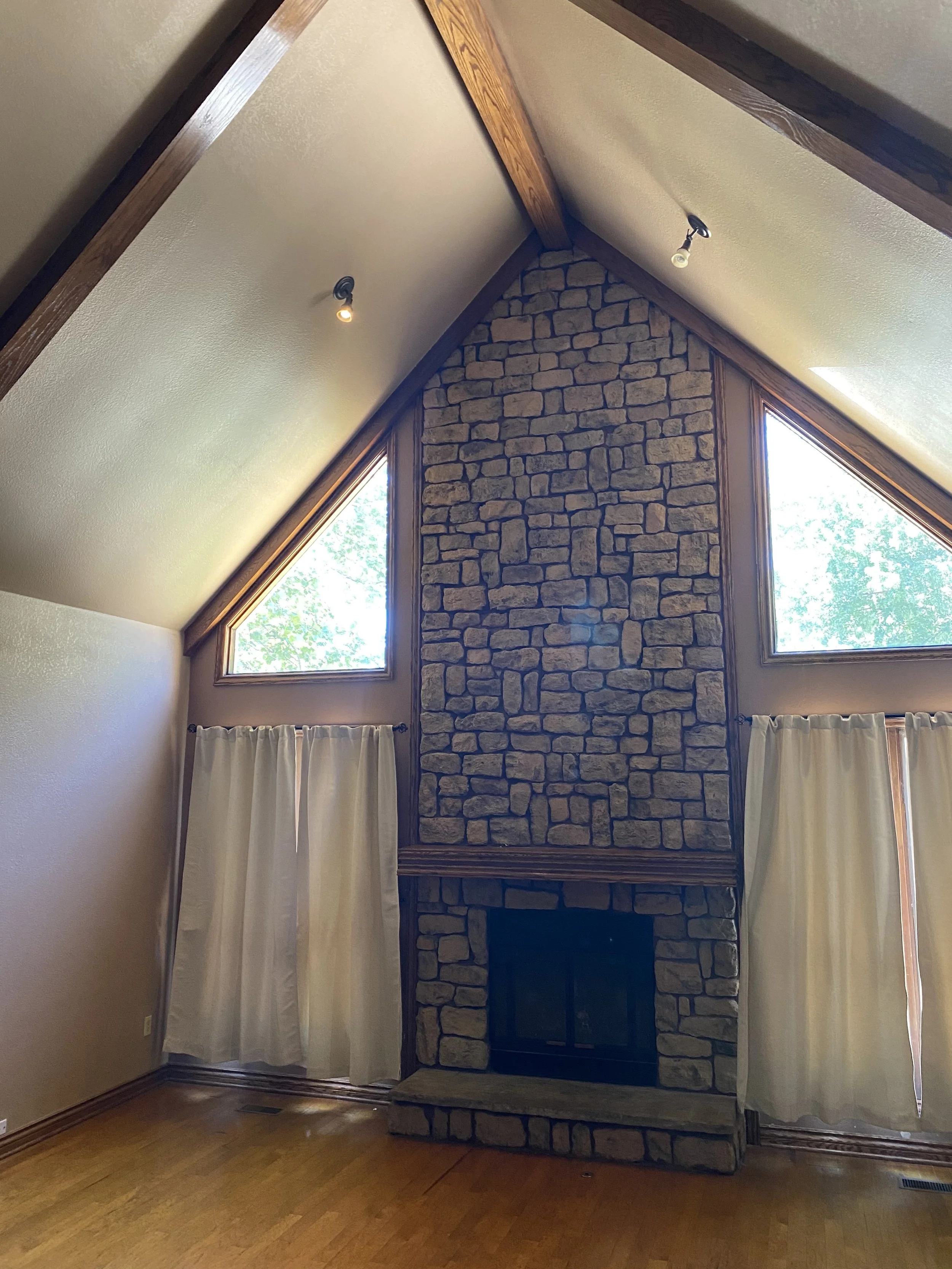 Living room with a stone fireplace flanked by two large windows with curtains, a vaulted ceiling with wooden beams, and wood flooring.