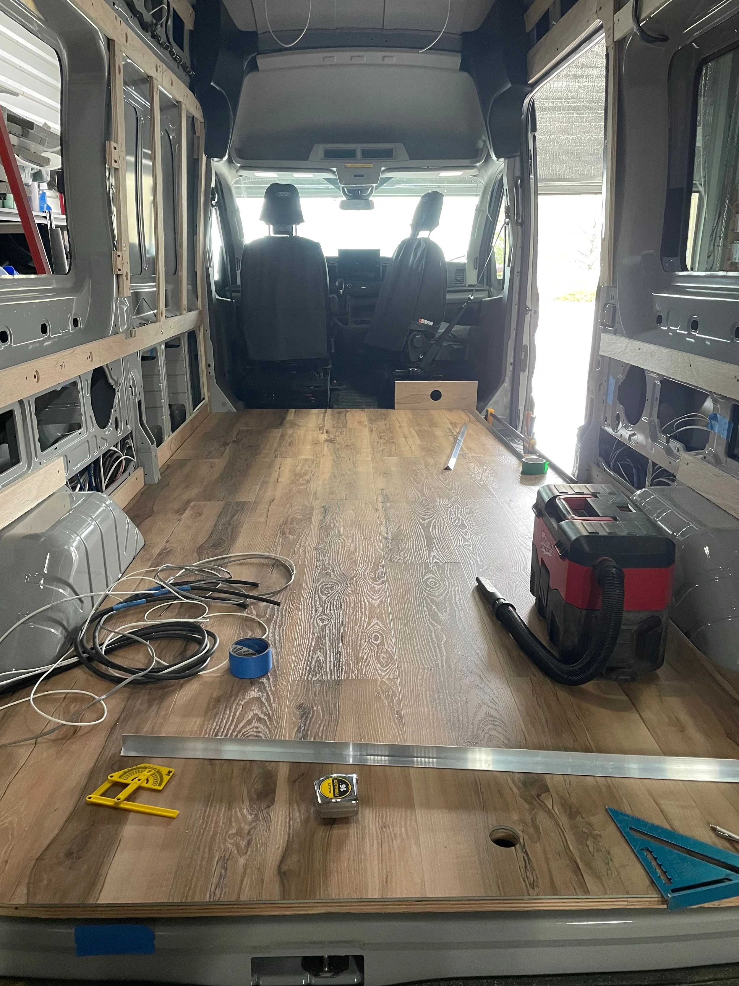 Inside the unfinished cargo area of a van being converted, with wooden flooring partially installed, tools, wires, and construction materials scattered around.