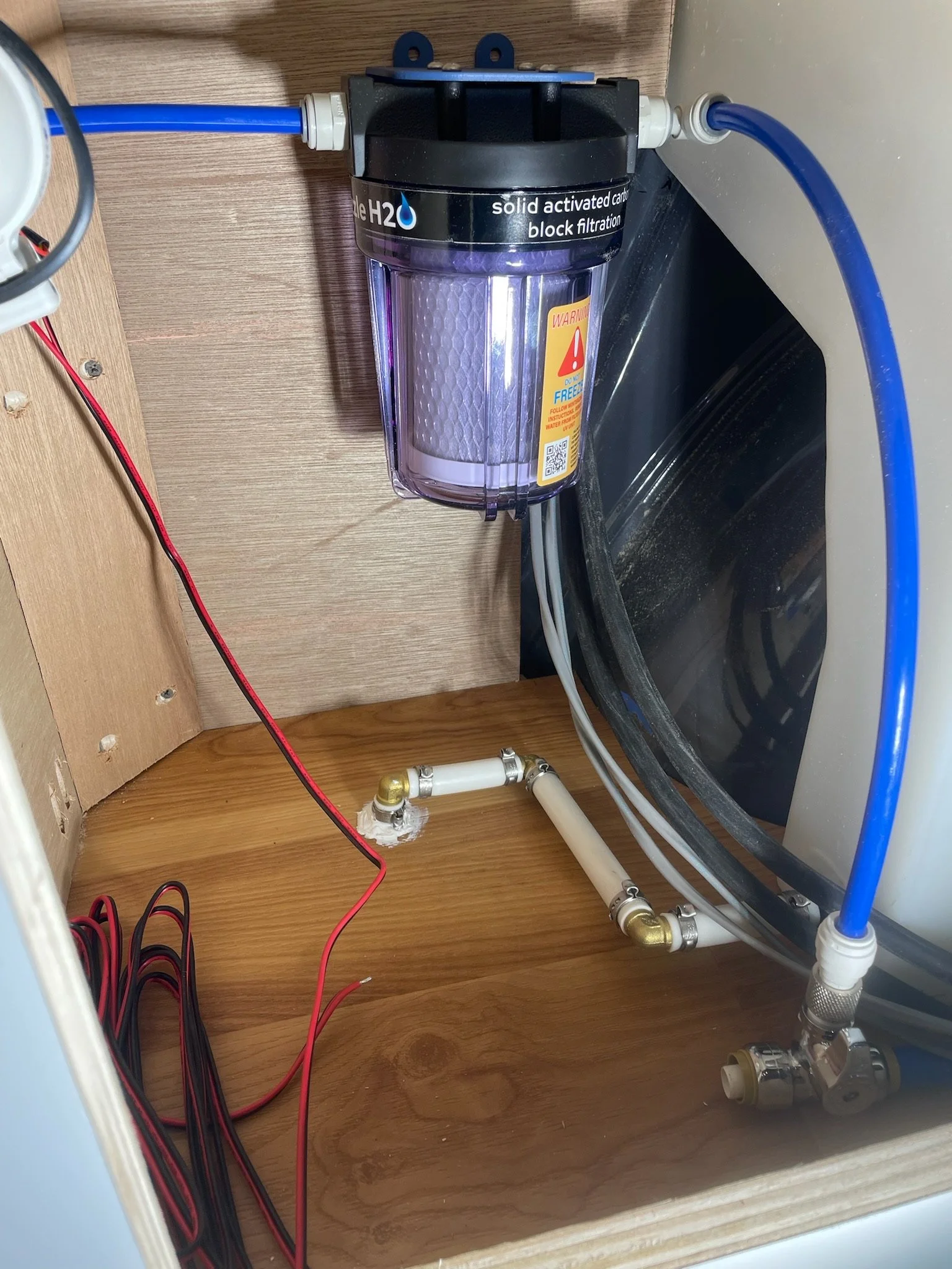 Under-sink water filtration system with blue and white tubing, red and black wires, and a wooden cabinet interior.