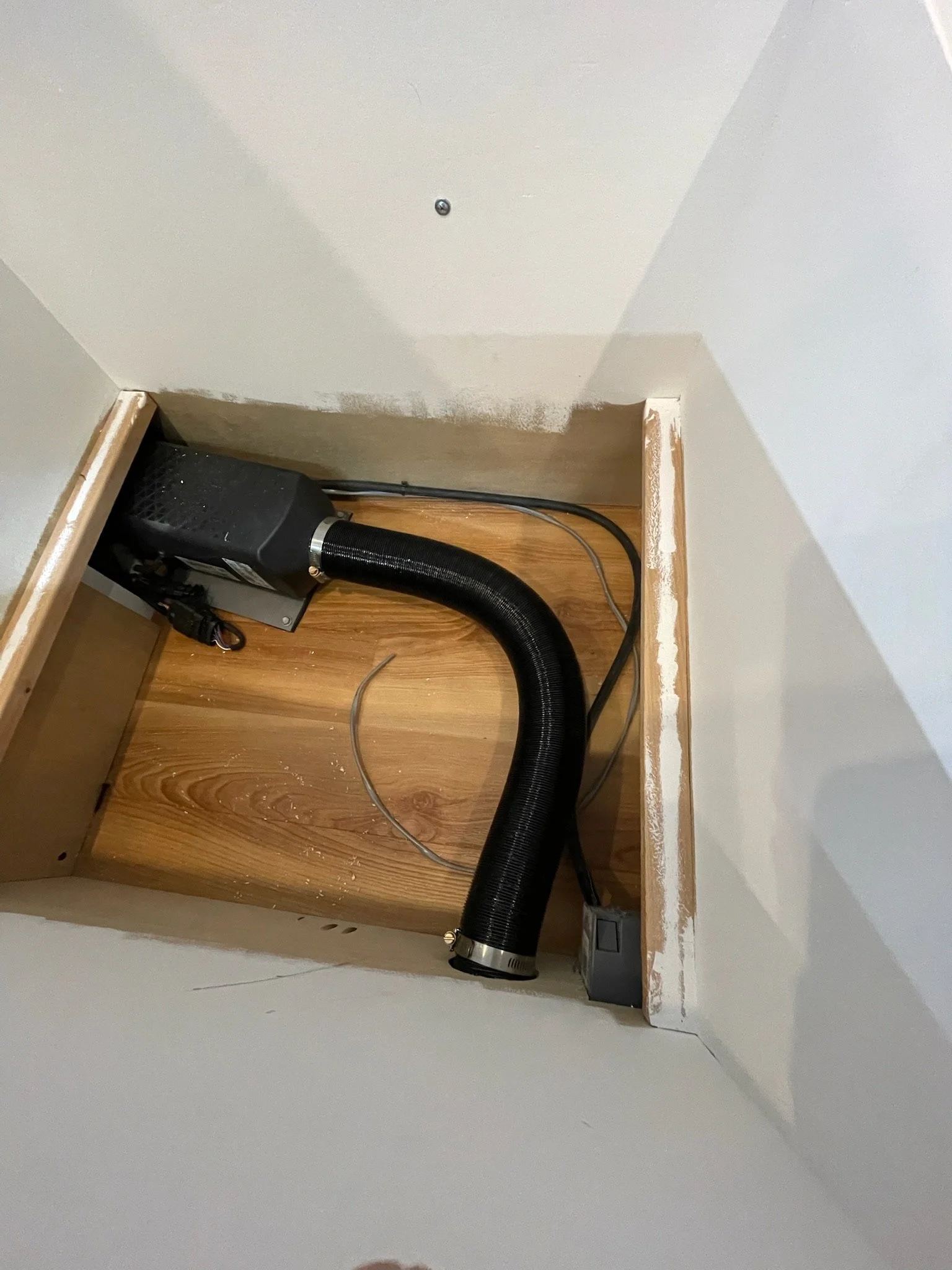 Interior space with a black flexible air duct pipe, a small heater or air blower unit, and a power outlet on a wooden floor.