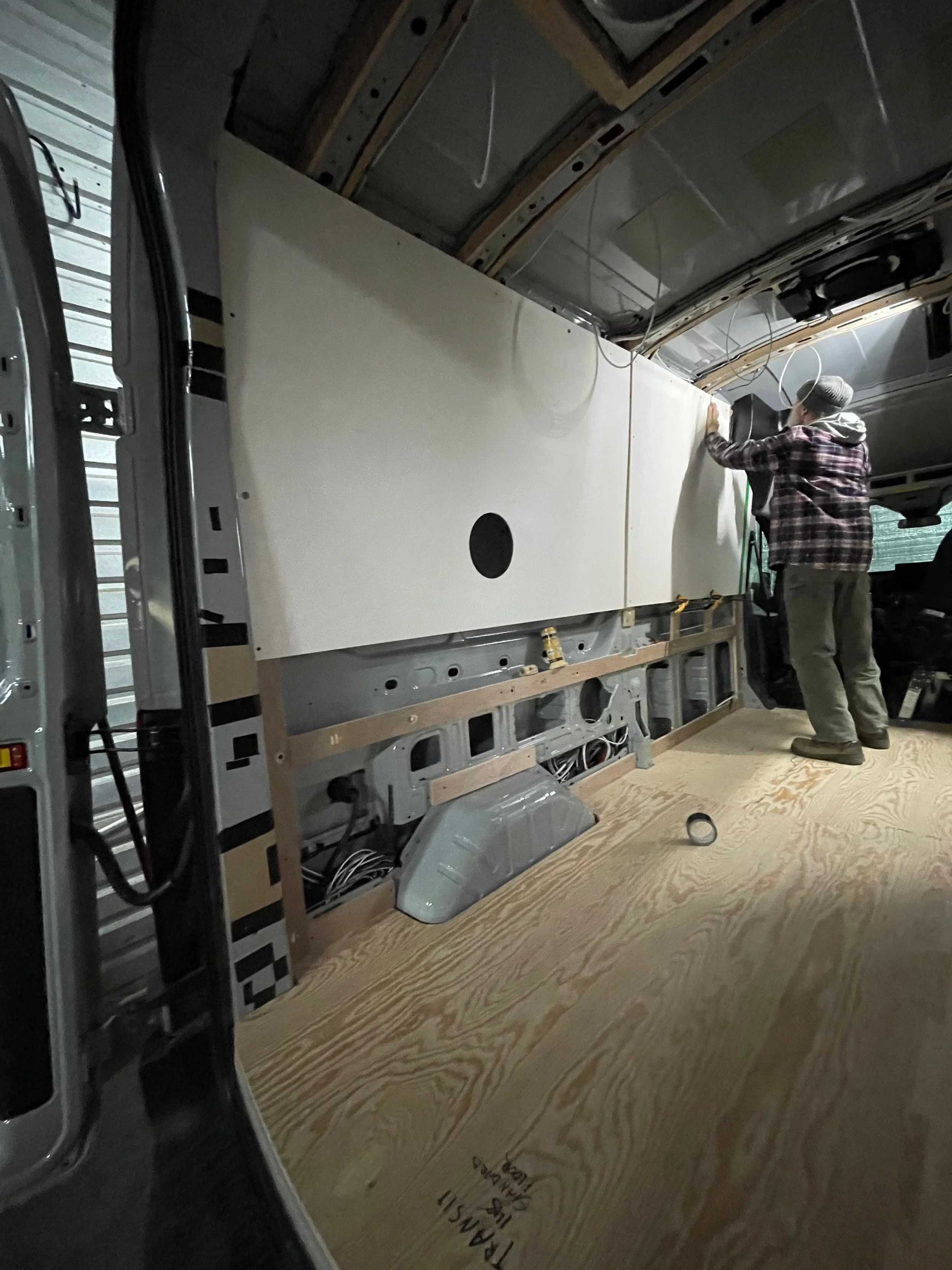 A person installing or working on a large white panel inside a commercial vehicle, possibly a van or RV, with exposed interior framing and wiring.