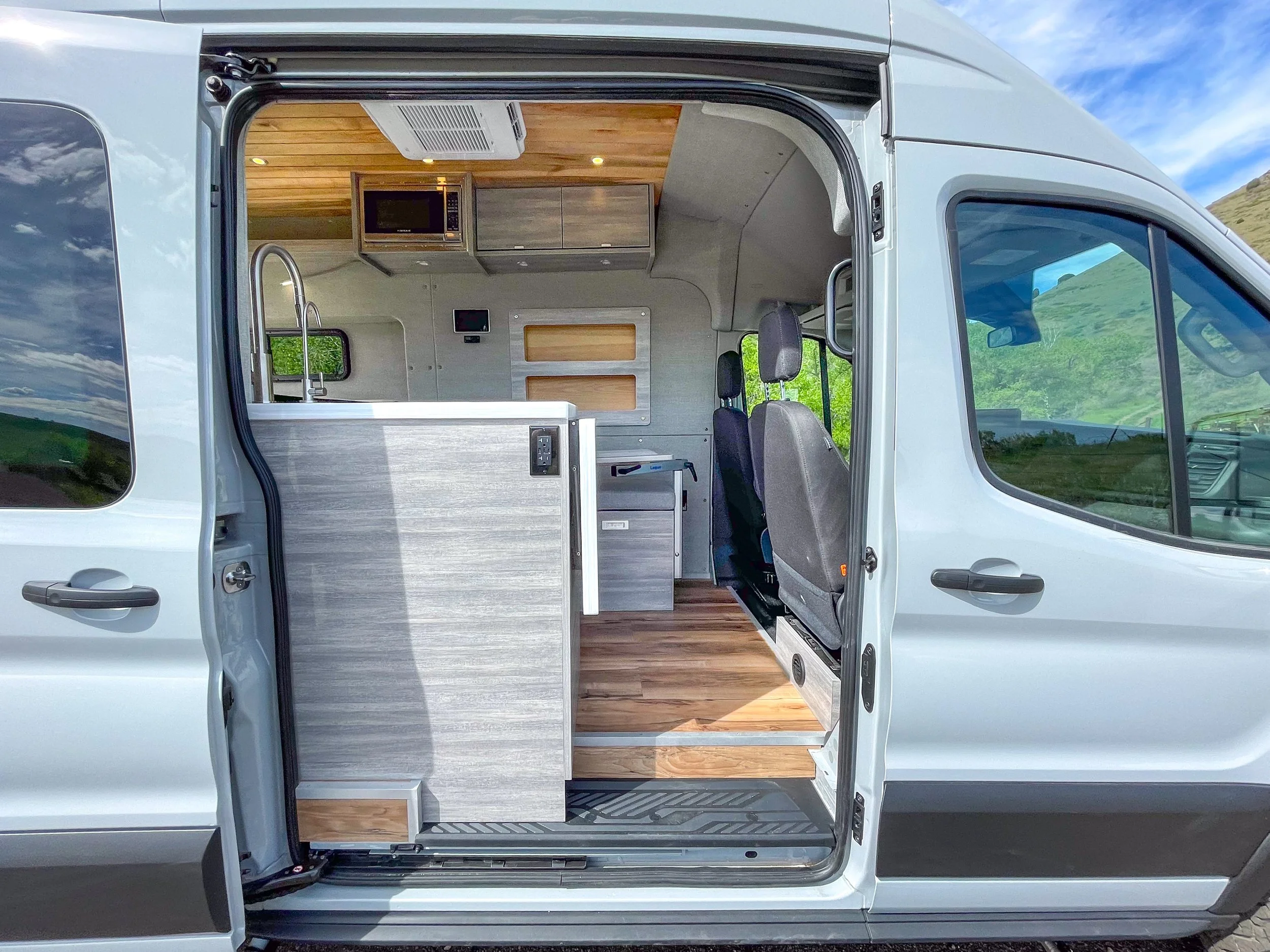 Interior view of a compact camper van with kitchen area, microwave, countertop, and seating, set against a background of green hills and blue sky.