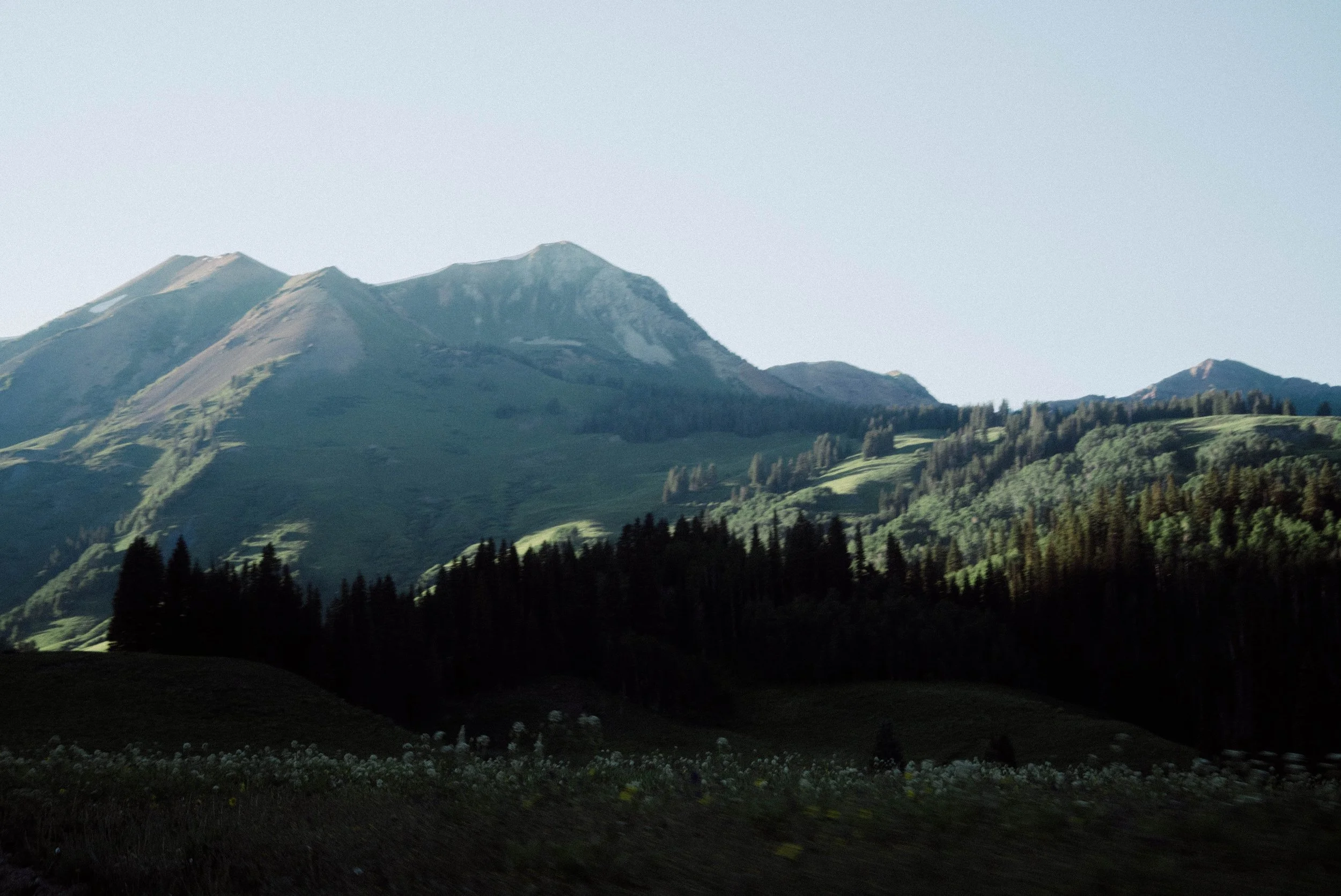 Mountain landscape with green hills and dense forest in foreground, clear sky above.