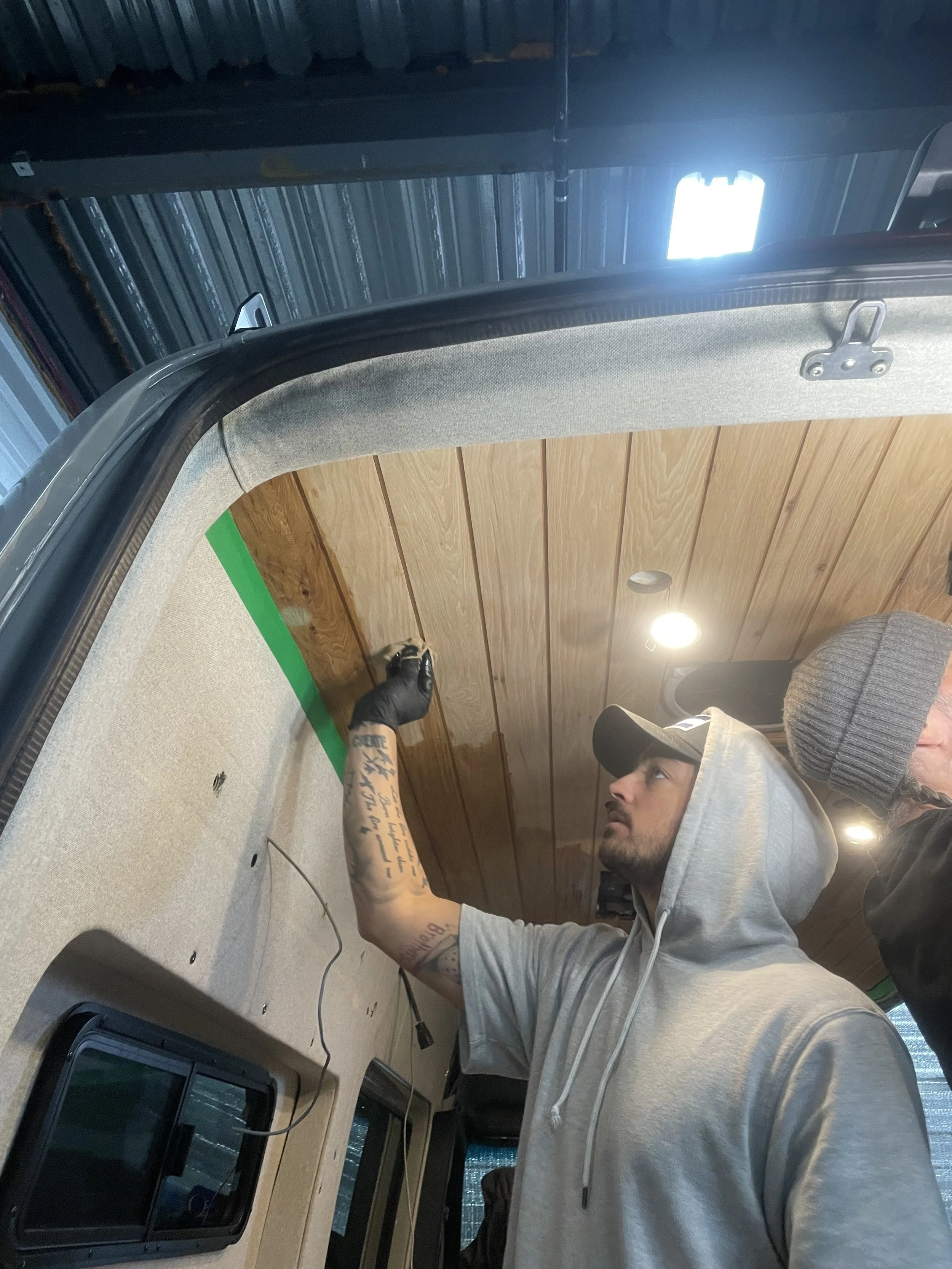 Two people working on the interior of a vehicle, installing wood paneling on the ceiling. One person is wearing a hoodie and cap using a tool, while the other is wearing a beanie.