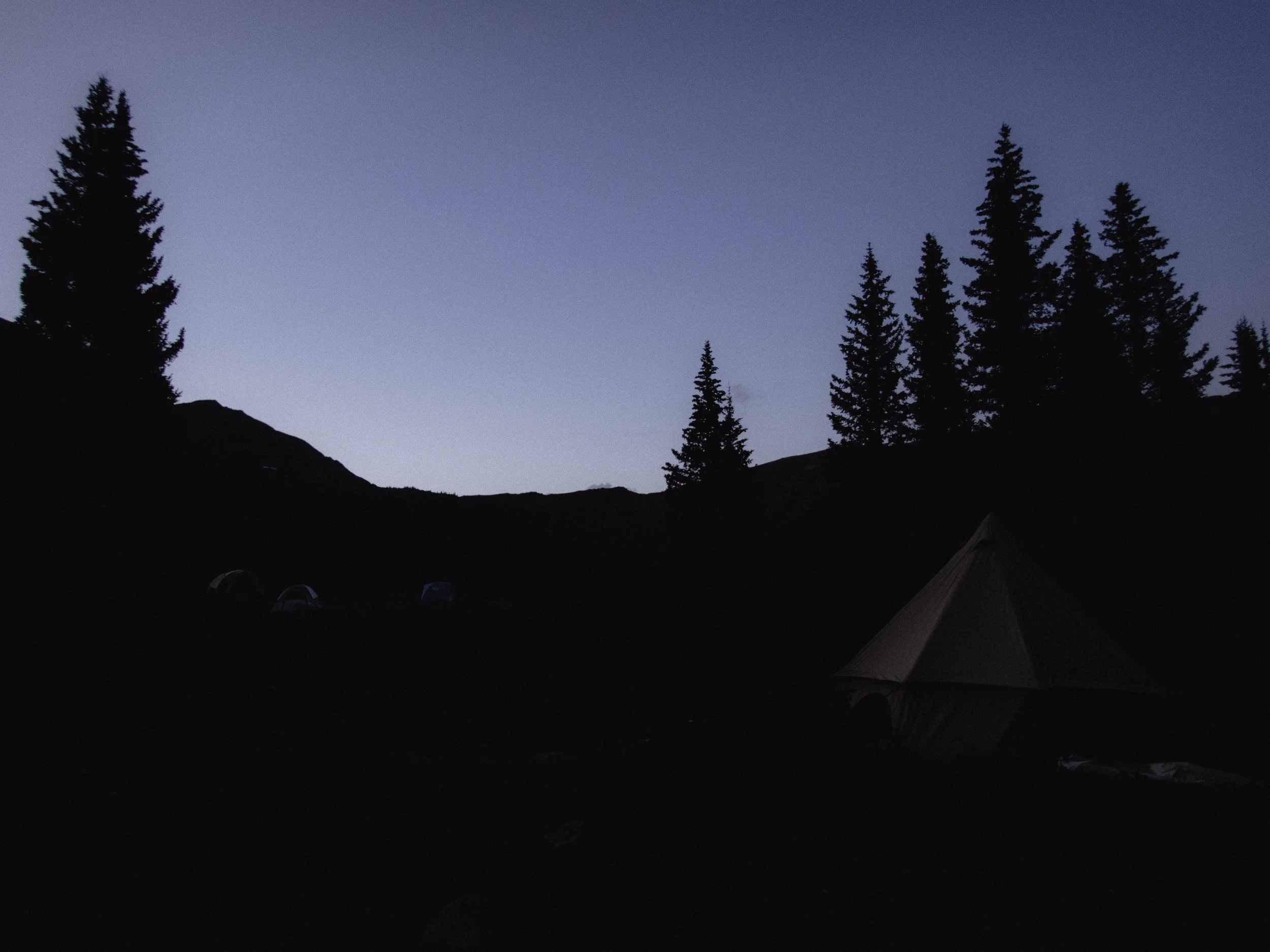 A dark silhouette of a camping area with tents and tall trees under a dusky sky at twilight.