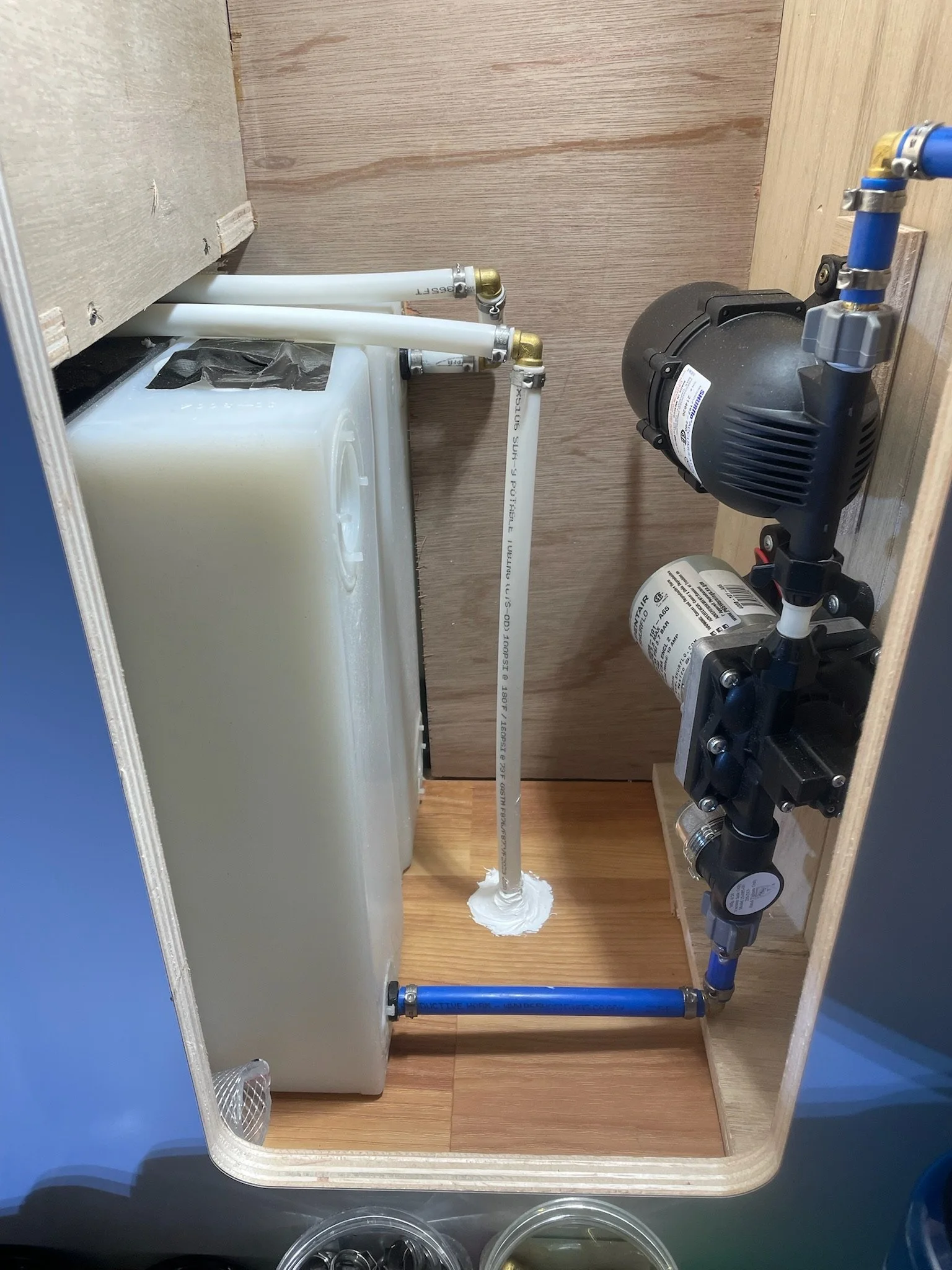 Inside view of a small storage compartment containing a white water tank, white and blue pipes, and a water pump.