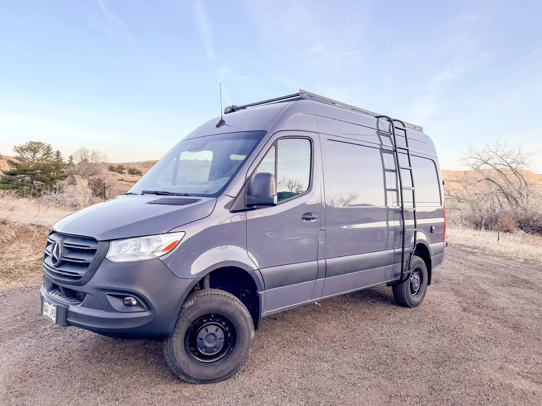 Gray camper van parked in a rural outdoor setting with dry grass and trees in the background.