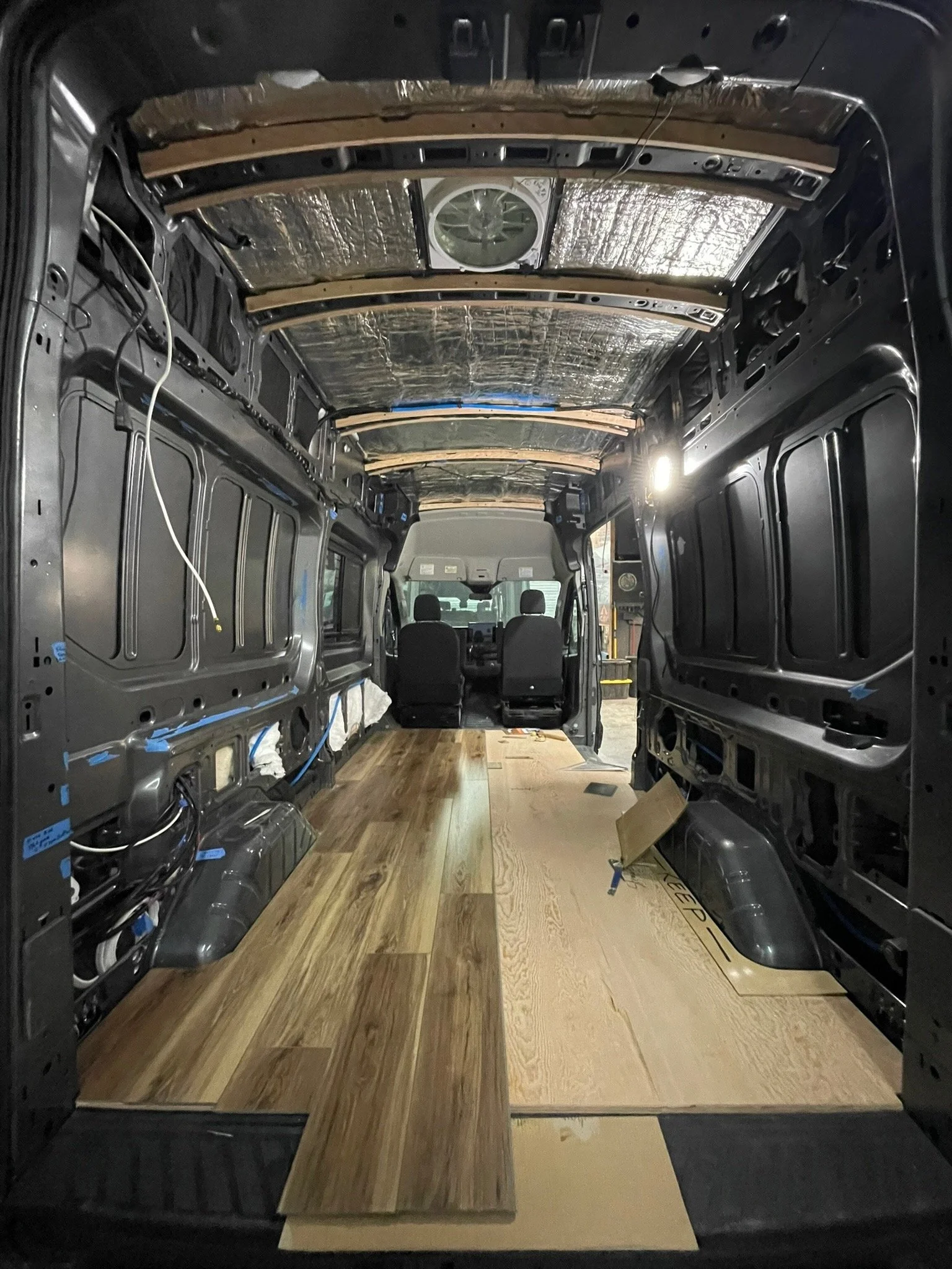 Interior of a vehicle van under conversion, with insulation and wooden flooring in progress, driver and passenger seats visible in the front.