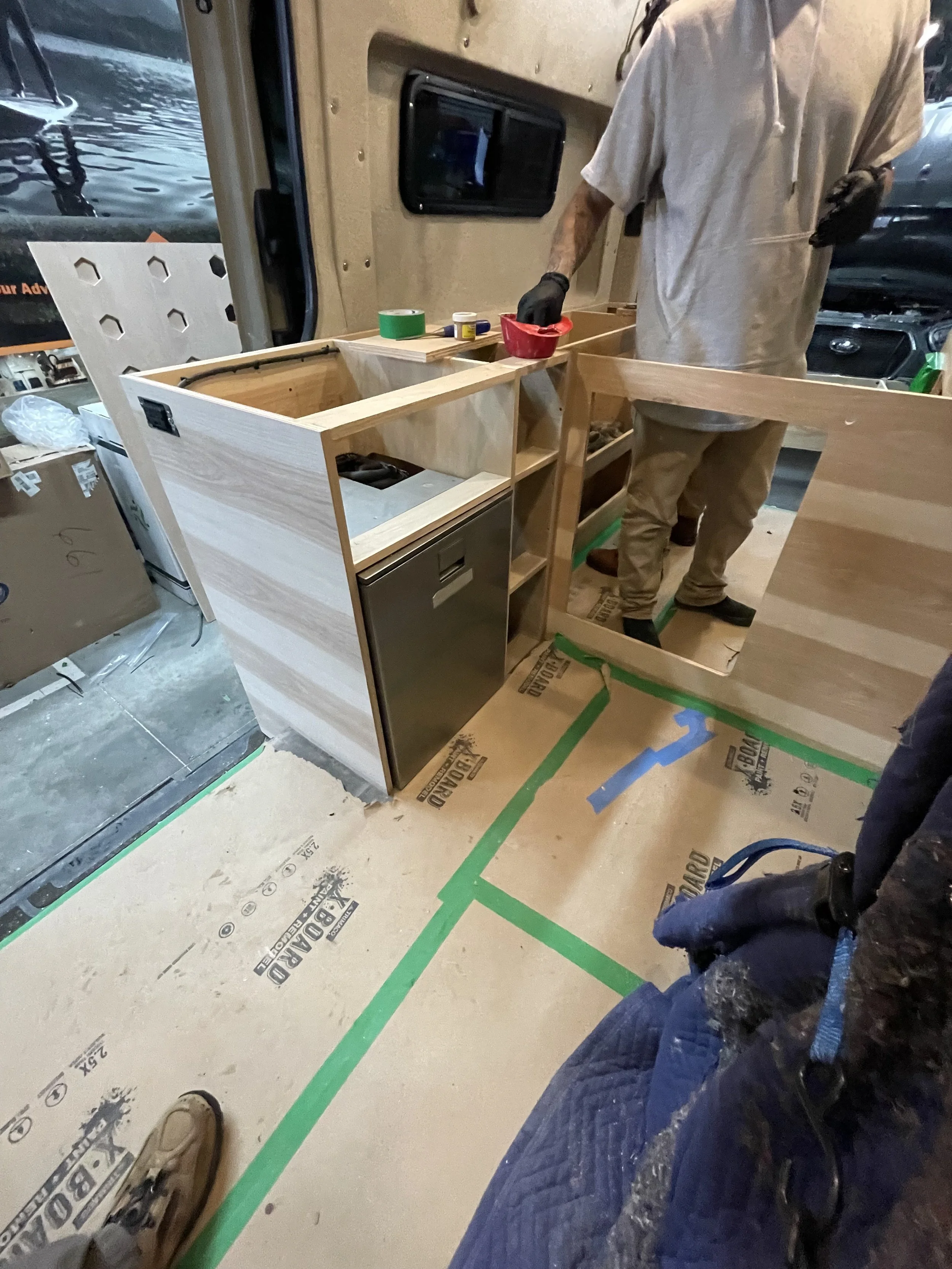 Van interior being converted with wooden cabinetry, a countertop, and insulating materials. A person is working on the construction, holding a red item and wearing gloves.