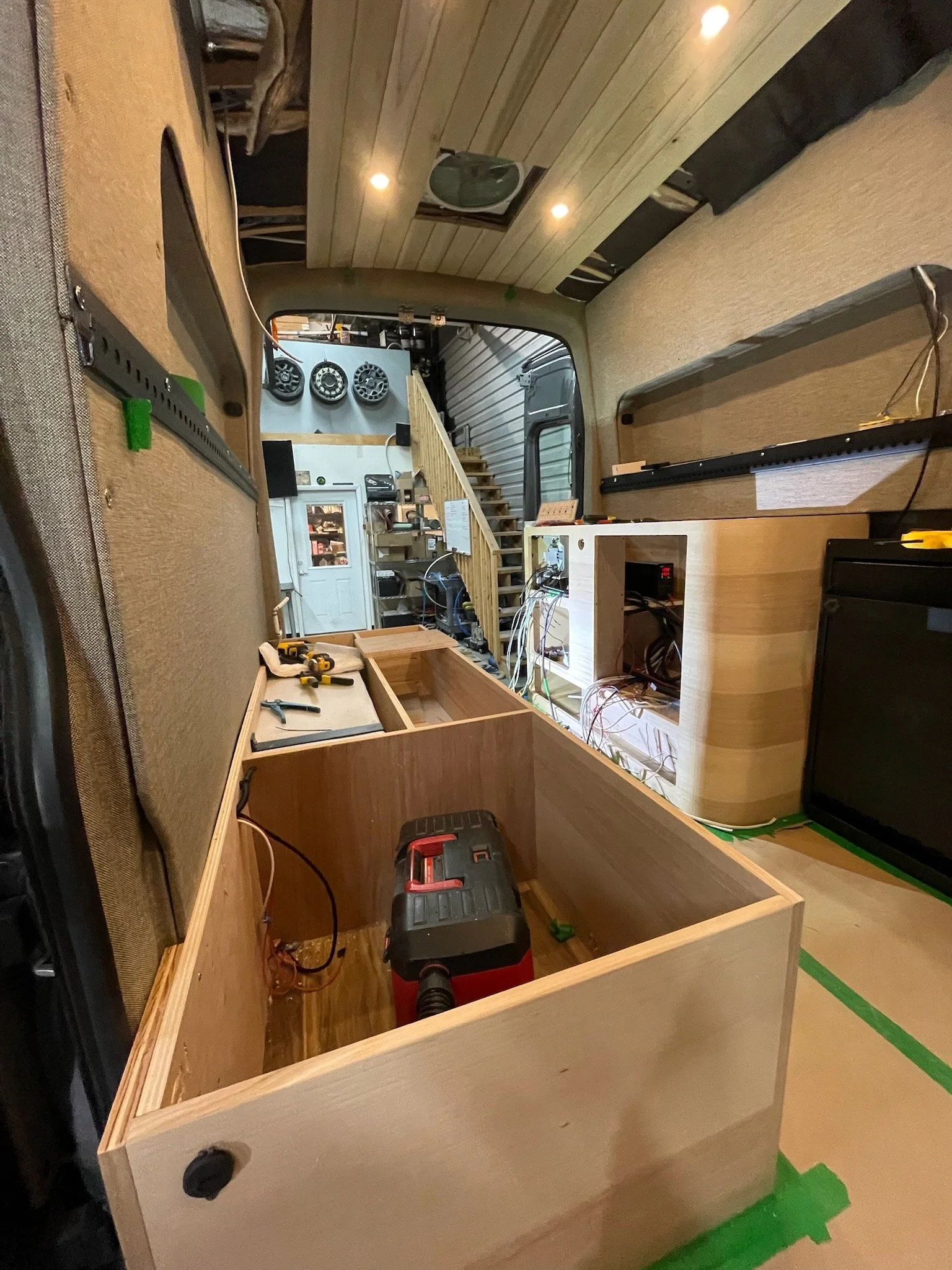 Interior of a small custom-built camper van under construction, showing unfinished wood cabinetry, tools, wiring, and a staircase leading to the upper level.