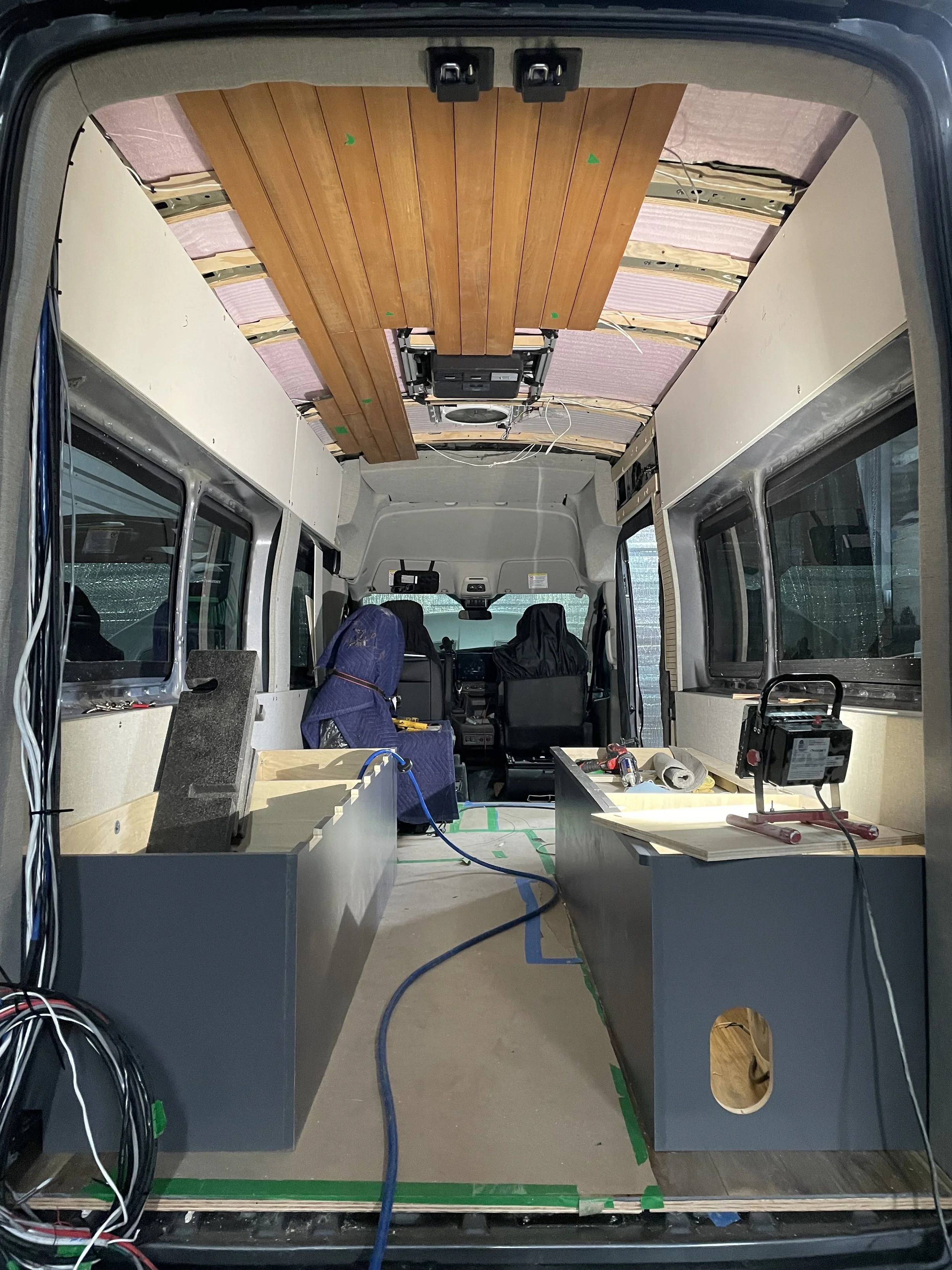Interior view of a van being converted into a camper with partially installed wood paneling on the ceiling, two storage units along the sides, and electrical wiring visible.