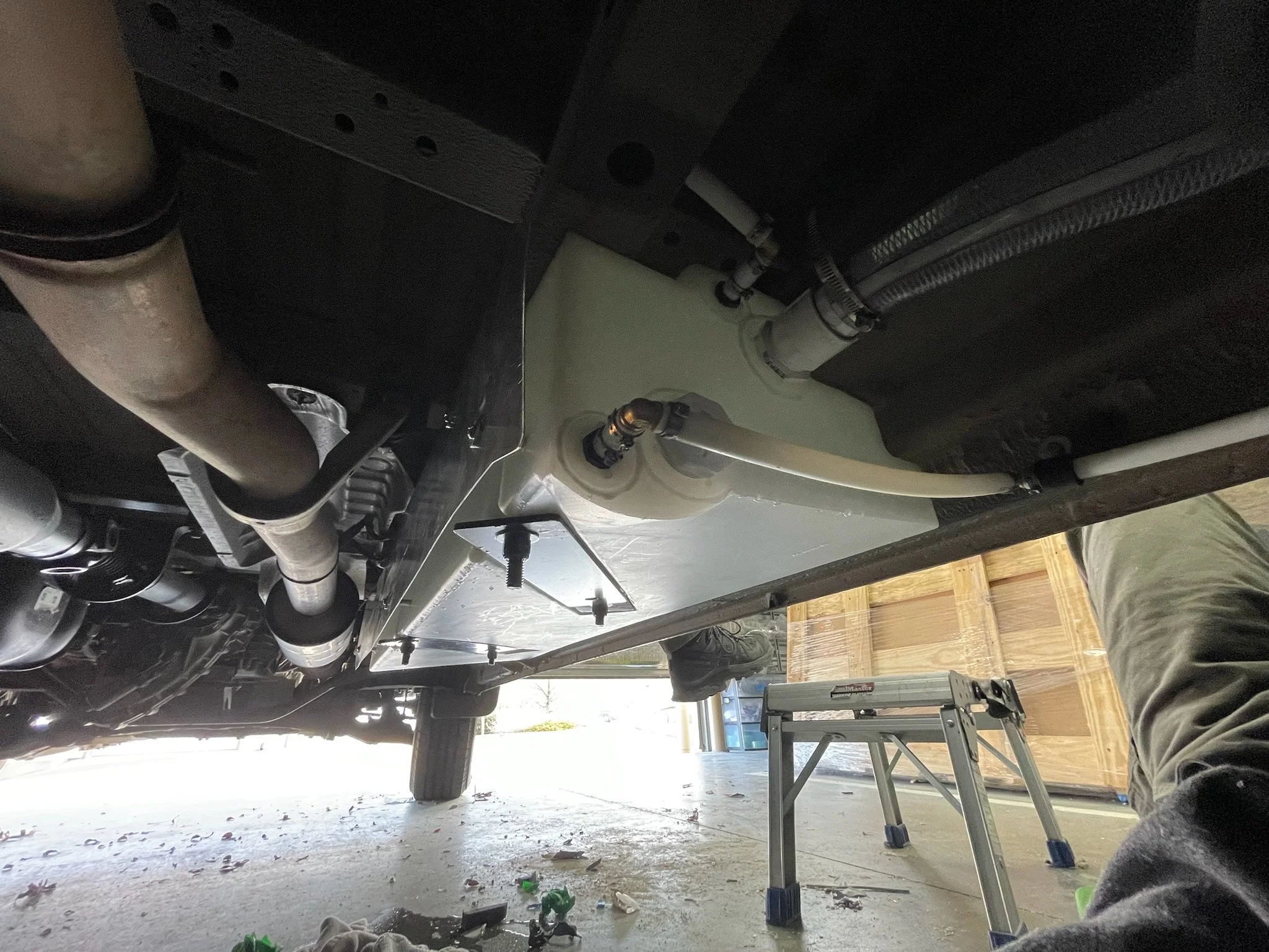 Underneath view of a vehicle showing exhaust system and storage tank with visible hoses and pipes. A person is working near the tank, with legs extended. A stepladder and garage setting are in the background.