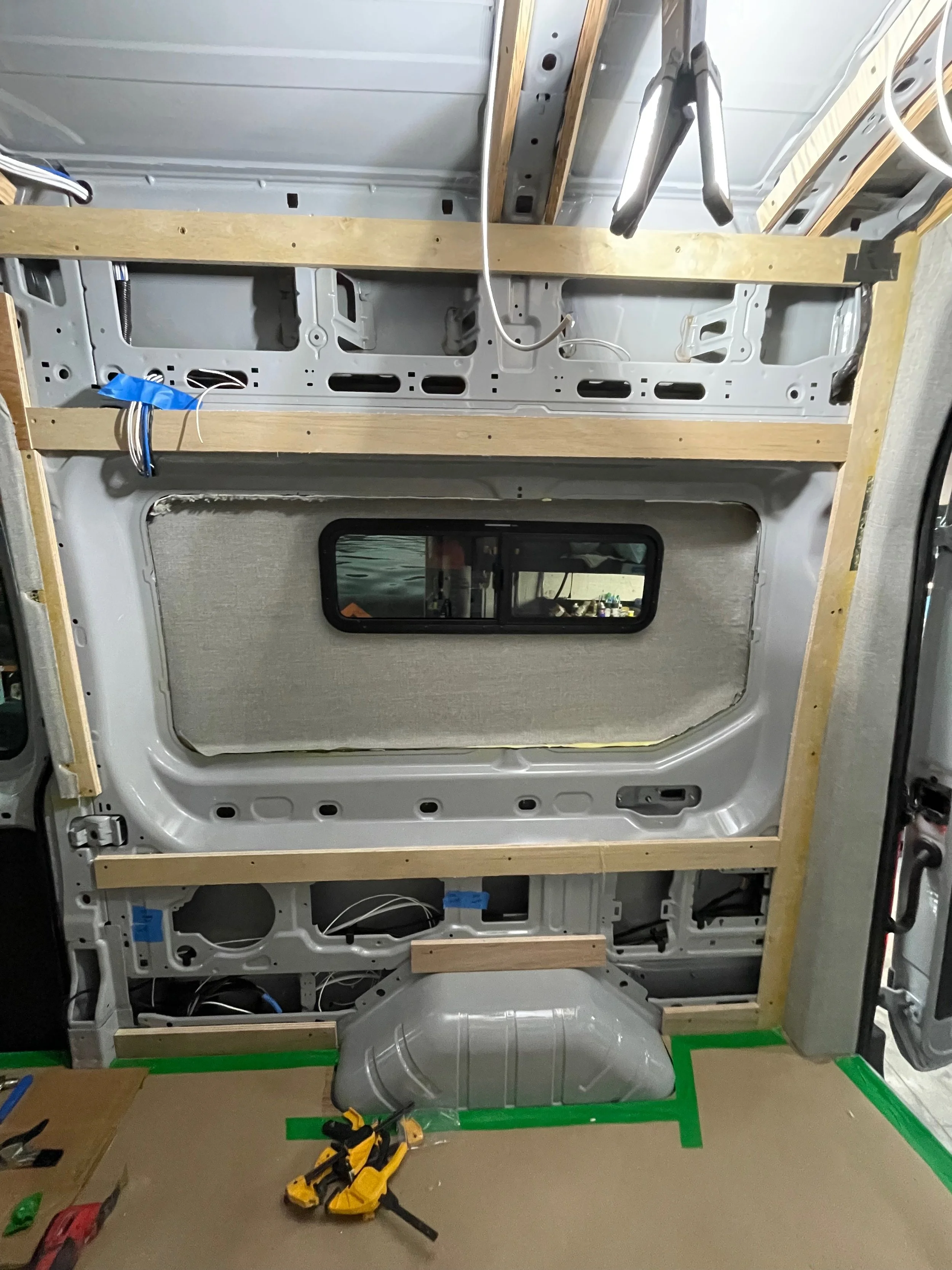 Interior of a van under renovation with exposed metal frame, wiring, and wooden supports, including a small side window.