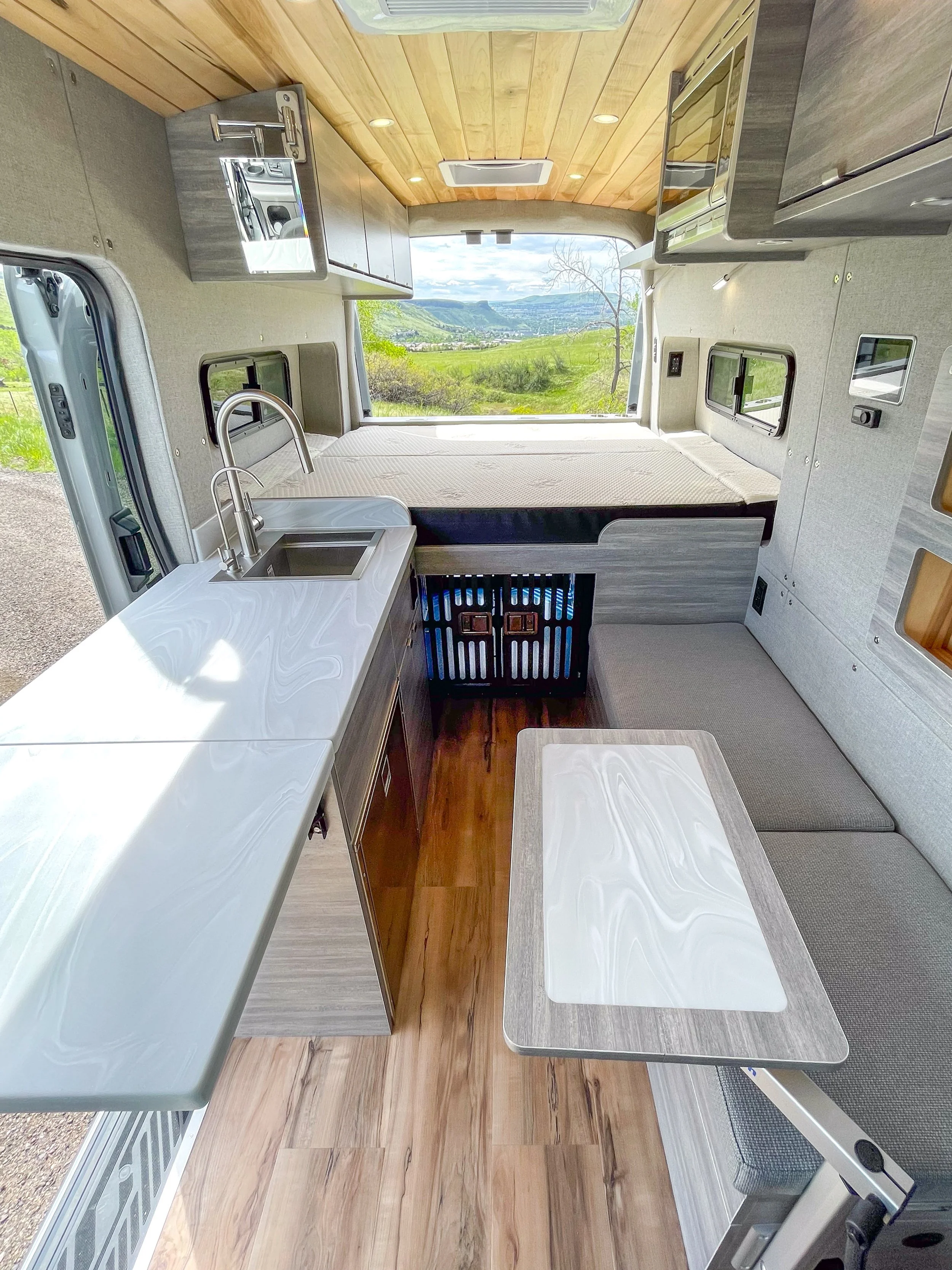 Interior of a camper van with a bed, a small kitchenette with a sink and countertop, a table, and a window showing a scenic outdoor view with hills and trees.