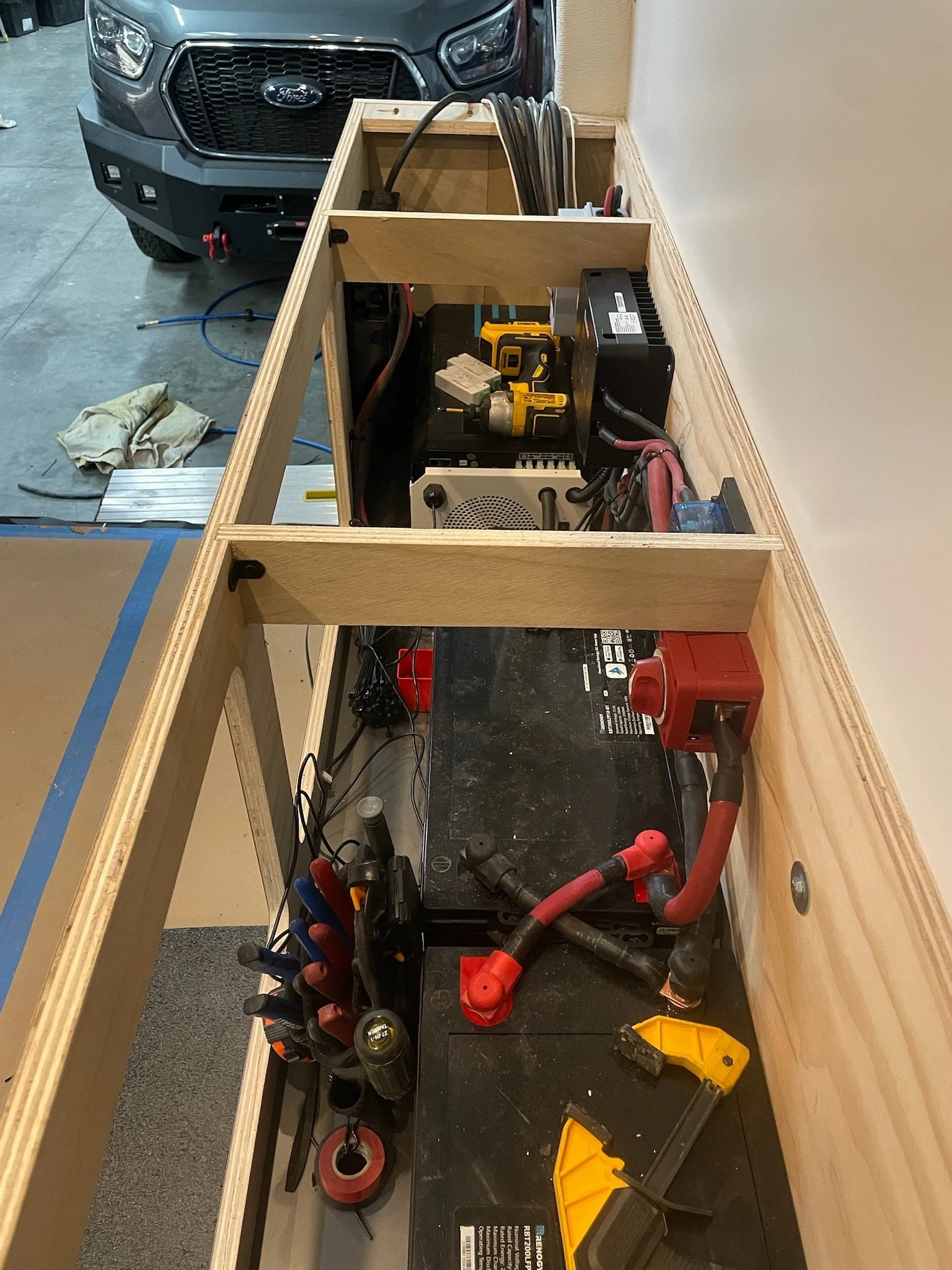 A wooden workbench with electrical tools and wires, a power drill, and a vehicle battery inside a garage with a Ford truck in the background.