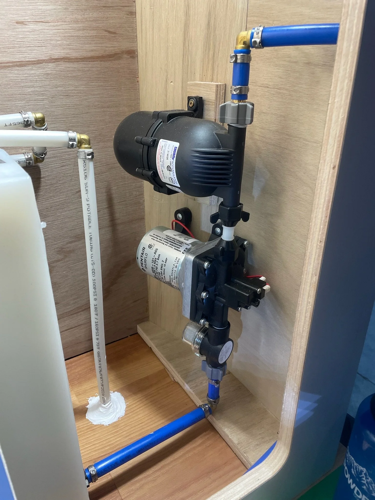 Water pump and plumbing system inside wooden enclosure with blue and white pipes.