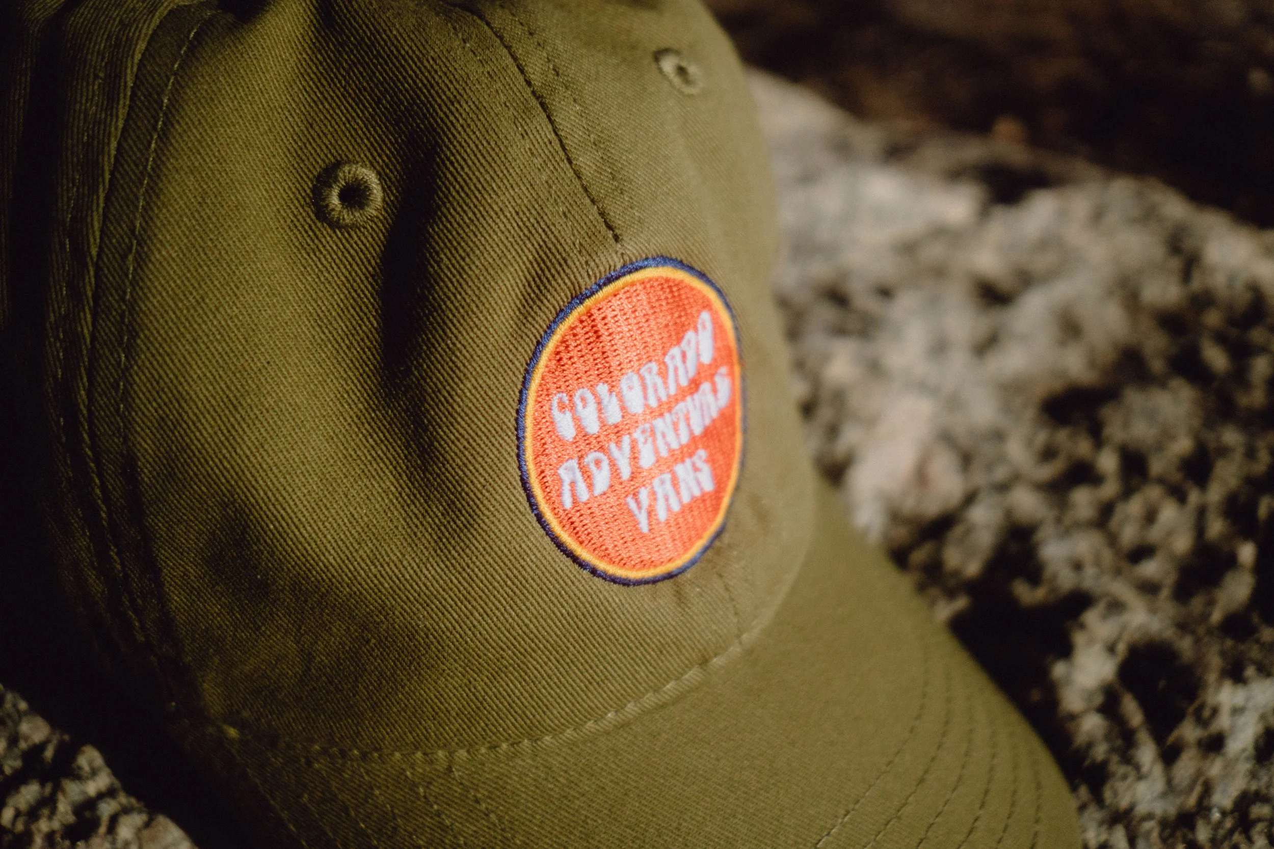 Close-up of an olive green cap with an orange circular patch that reads 'Colorado Adventure Vans'.