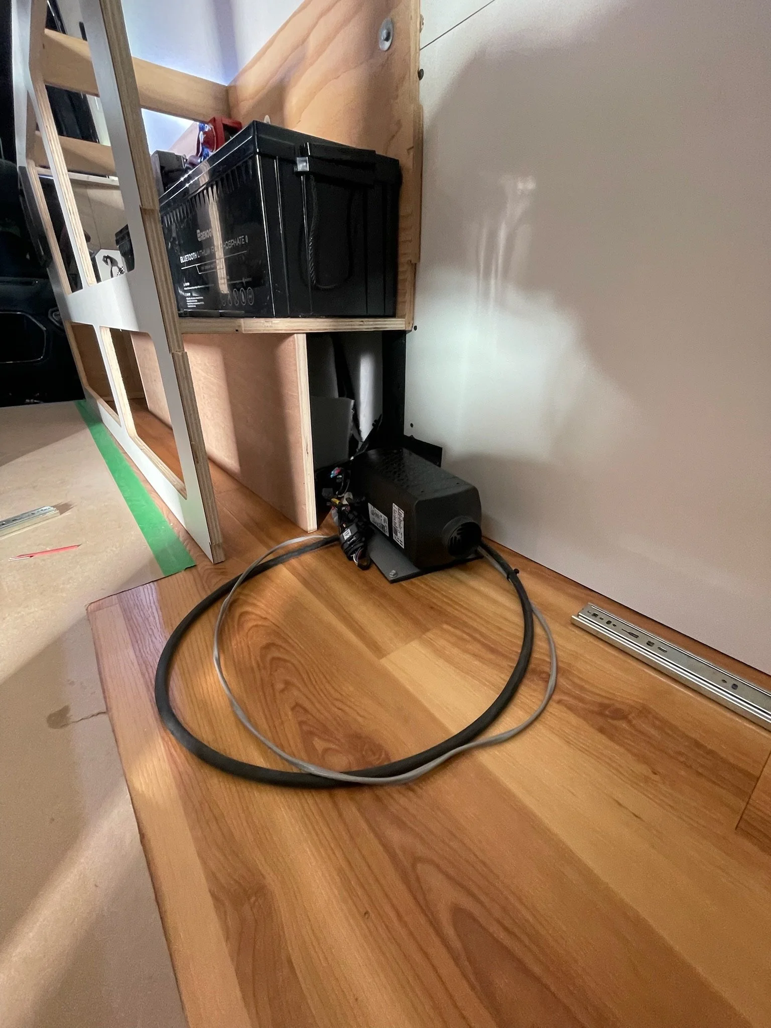 Interior of a van conversion with a wooden floor, a battery for power storage, electrical components, and wiring.
