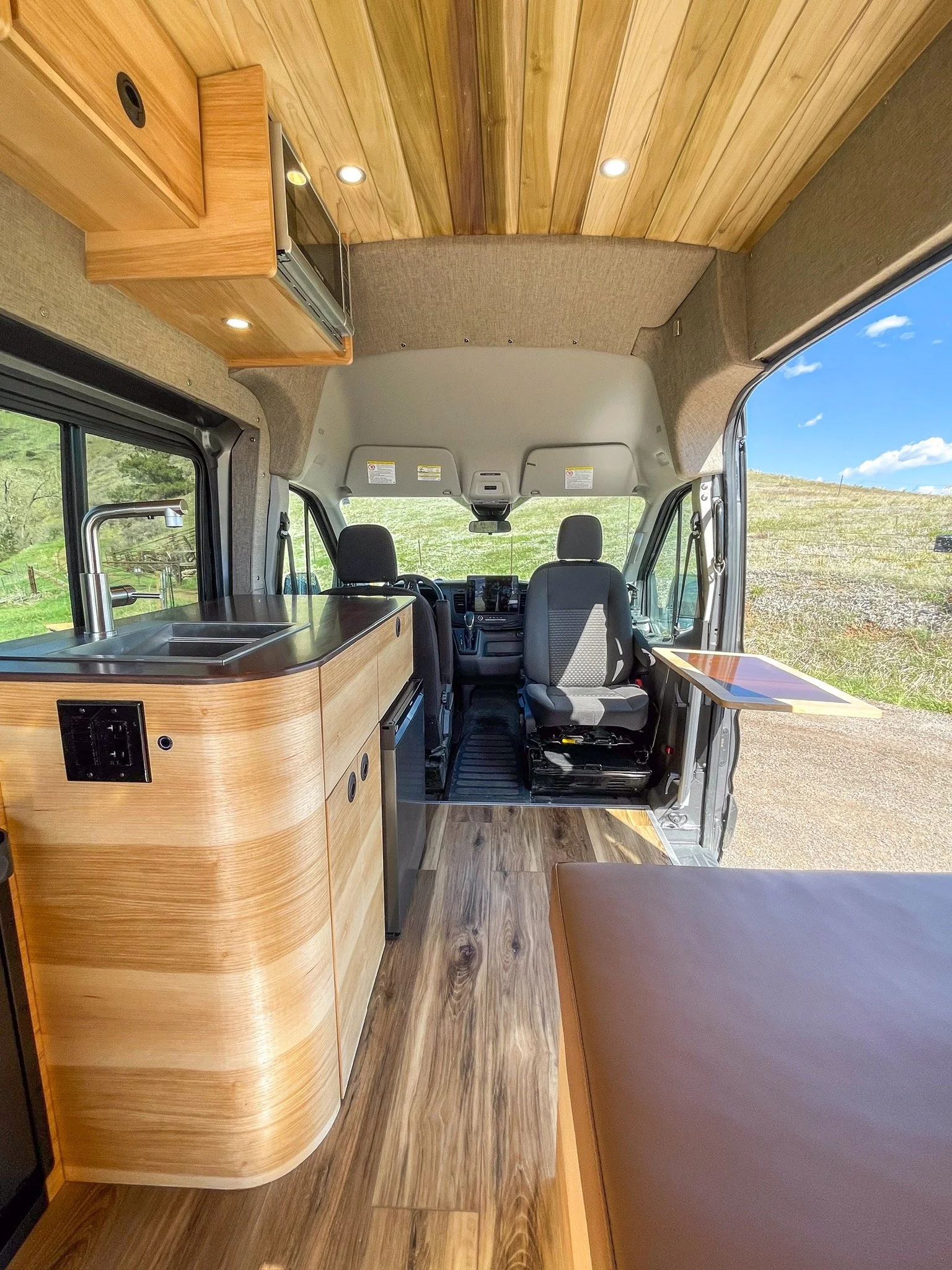 Interior view of a camper van with wood paneling, a countertop with a sink, a mounted microwave, two front seats, and a small outdoor table attached to the side.