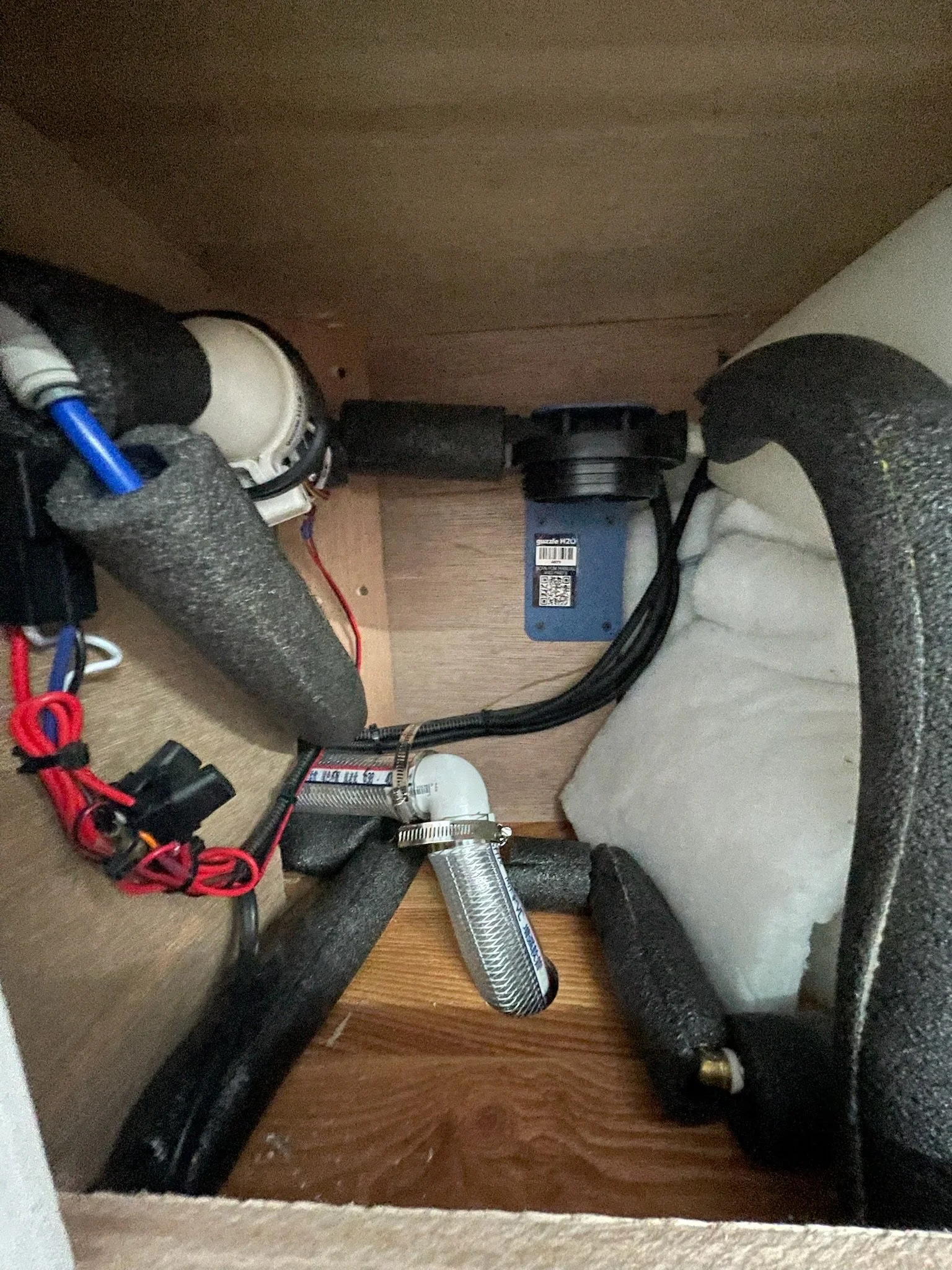 Plumbing system setup with pipes, insulation, wires, and a small pump inside a wooden enclosure.