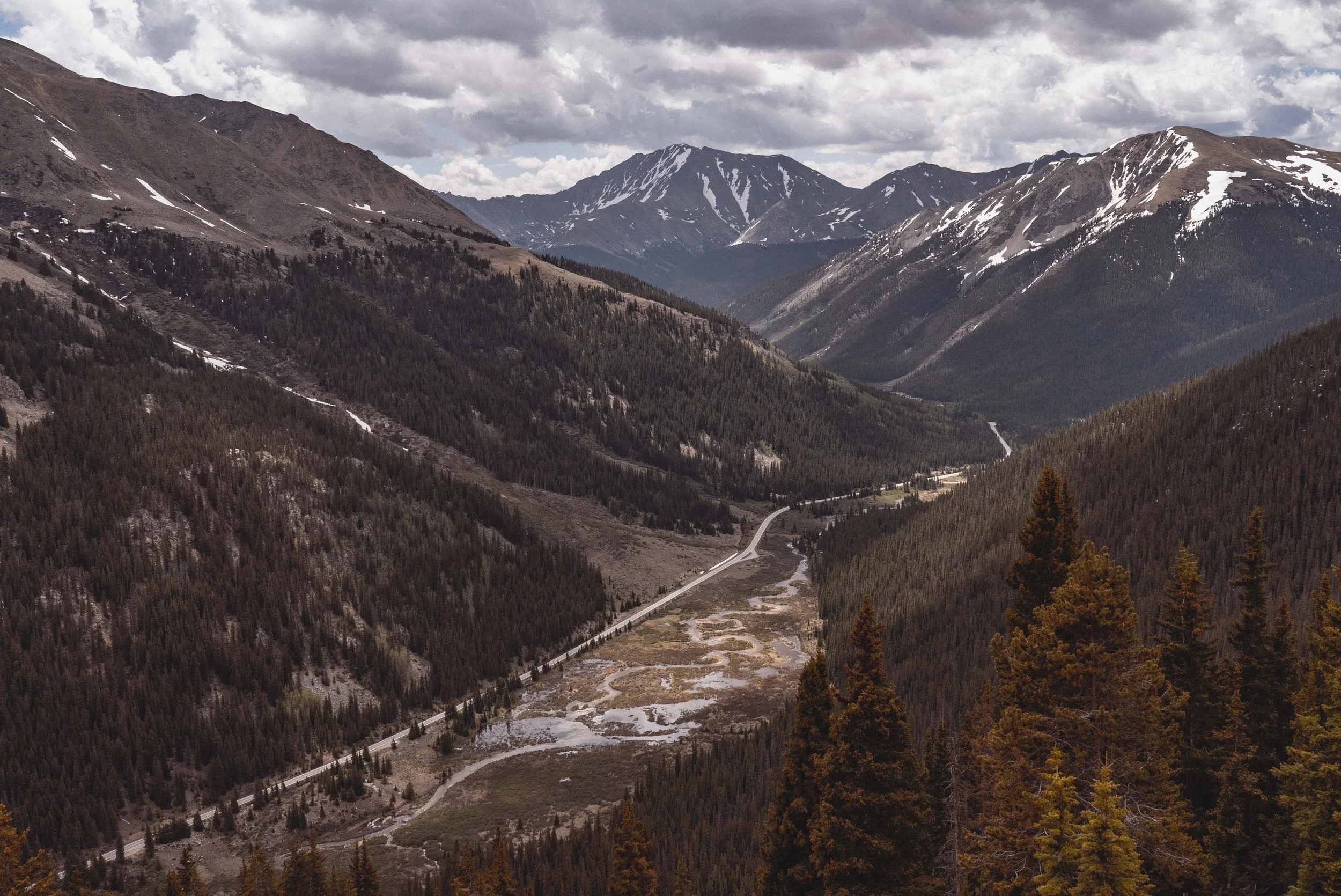 Mountain landscape with a winding road and a small stream in a valley, surrounding forested slopes, and snow-capped peaks under cloudy skies.