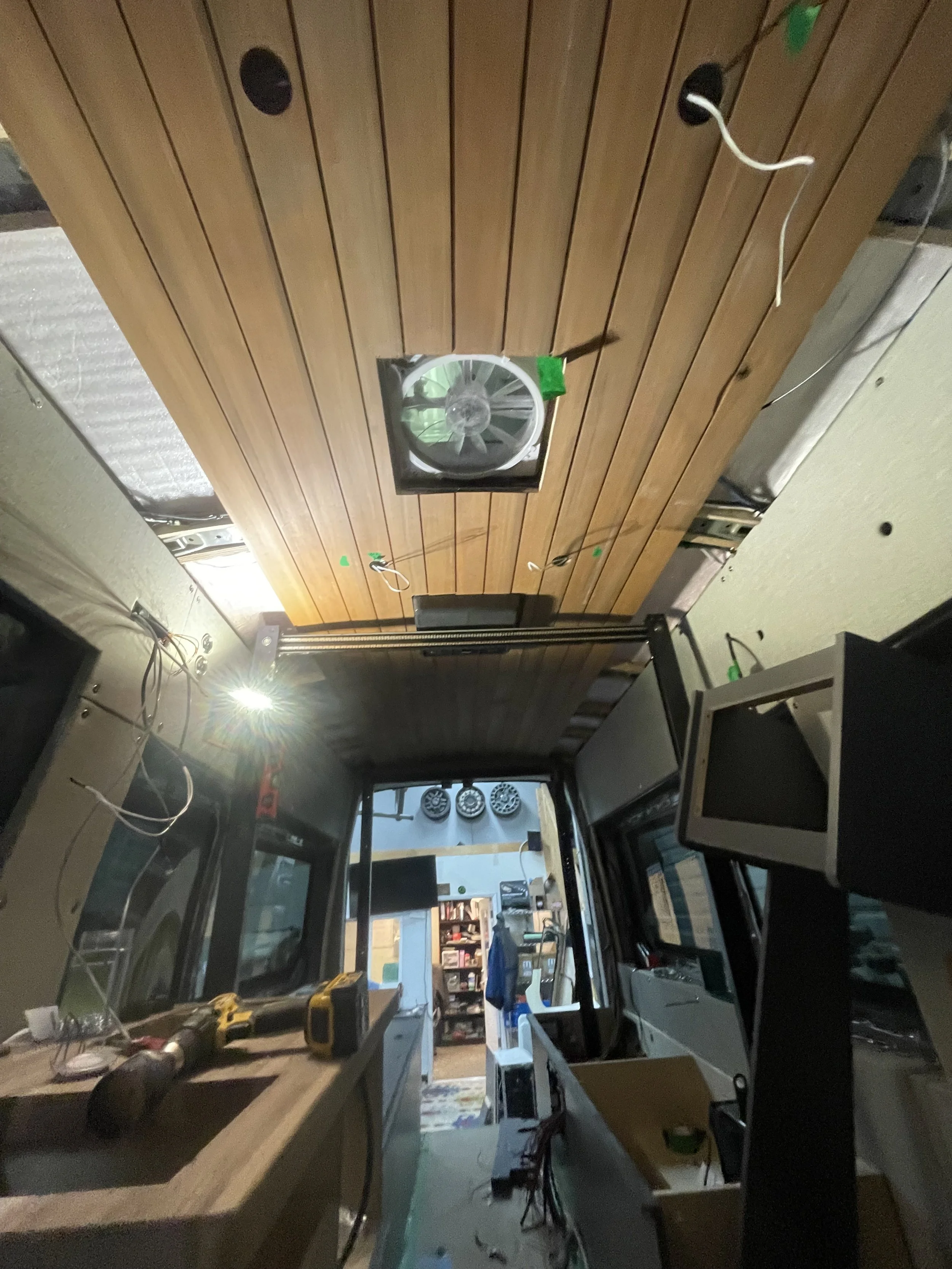 Interior of a partially constructed camper van with a wooden ceiling and built-in ventilation fan. Tools and materials are scattered on the floor and surfaces, indicating ongoing renovation work.