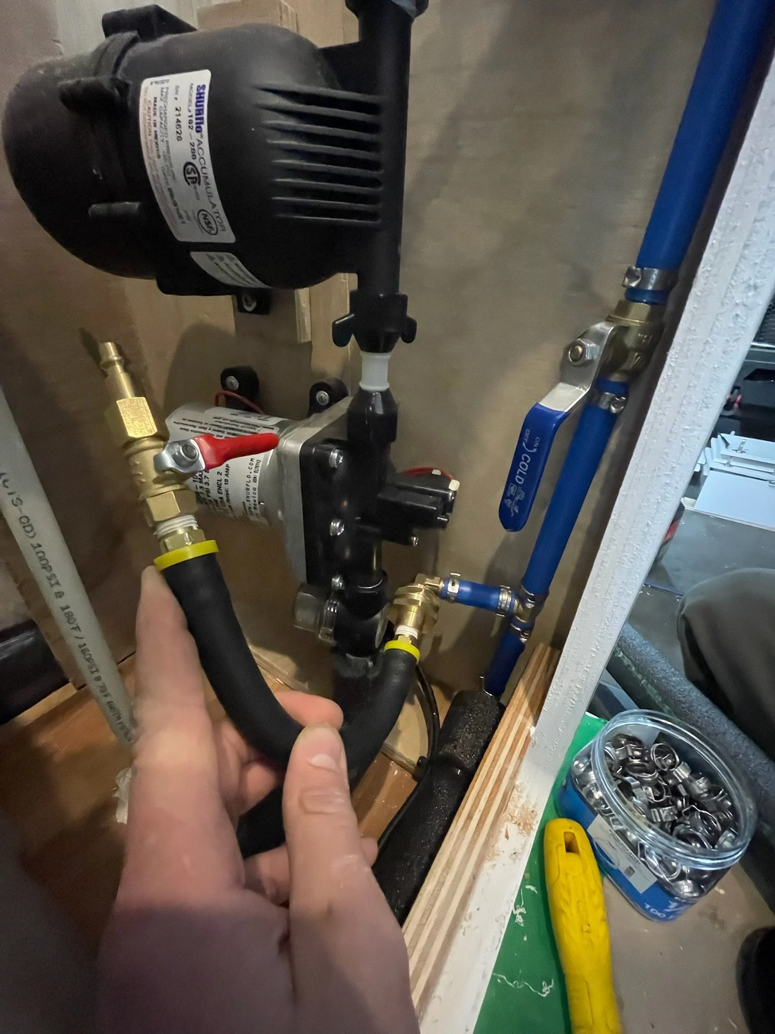Person holding black rubber hose near plumbing system with various pipes, valves, and a small pump.
