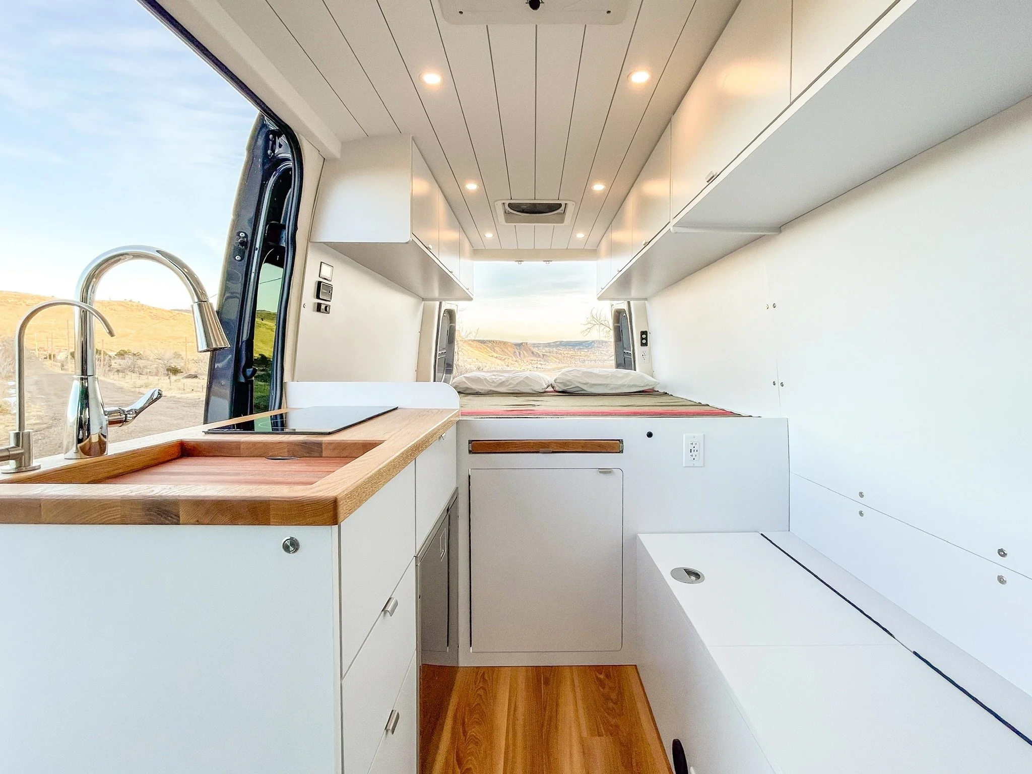 Interior of a modern camper van with a kitchen setup, including a sink with a faucet, wooden countertop, cabinets, and a bed in the background. The design features white cabinetry and wood flooring, with exterior scenery visible through an open door.
