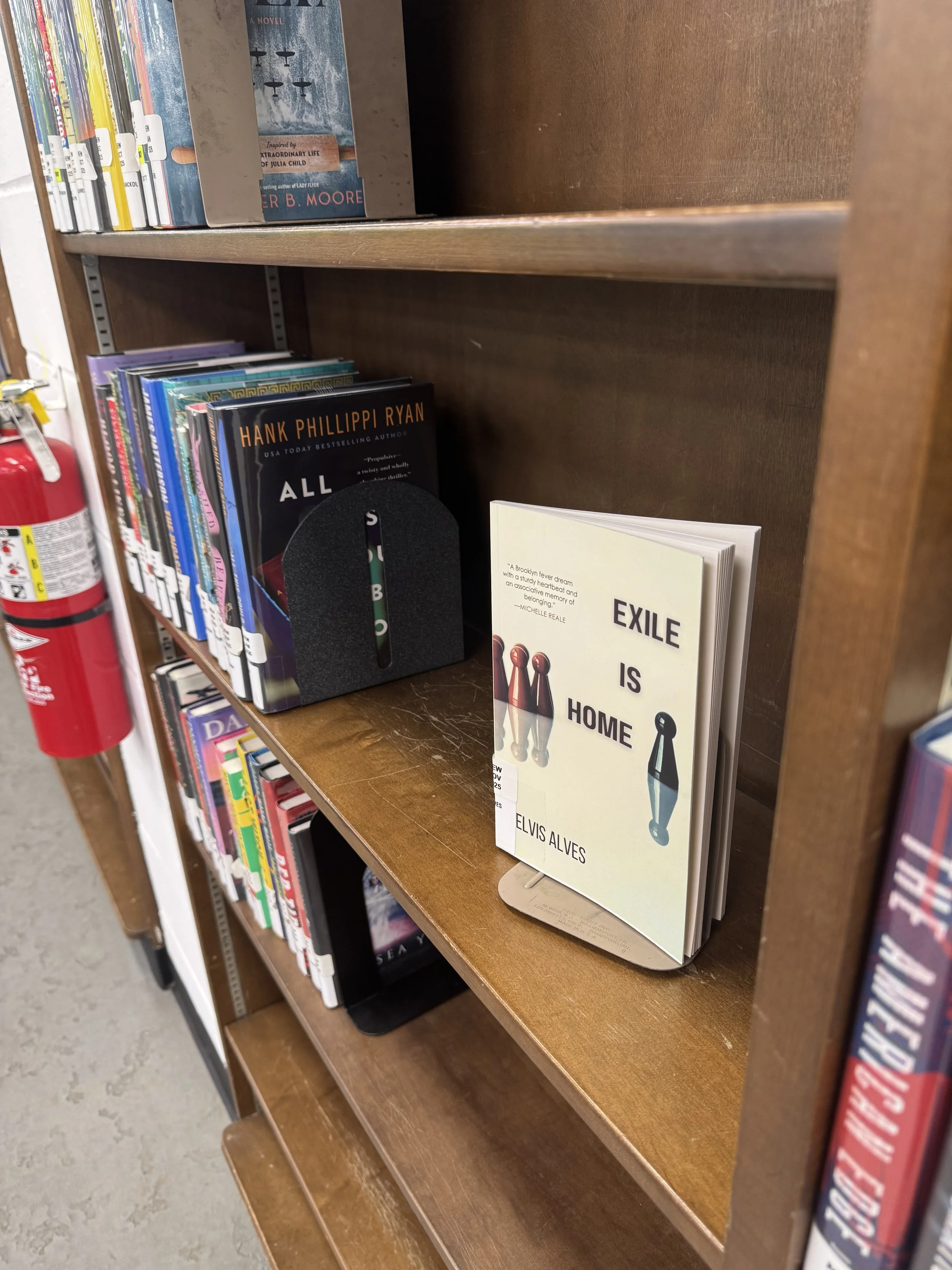 Library bookshelf with books and a small stand for a book titled 'Exile Is Home' by Elvis Alves. Other books include 'All the Light We Cannot See' by Anthony Doerr and a book by Hank Phillippi Ryan. A fire extinguisher is visible on the left side.