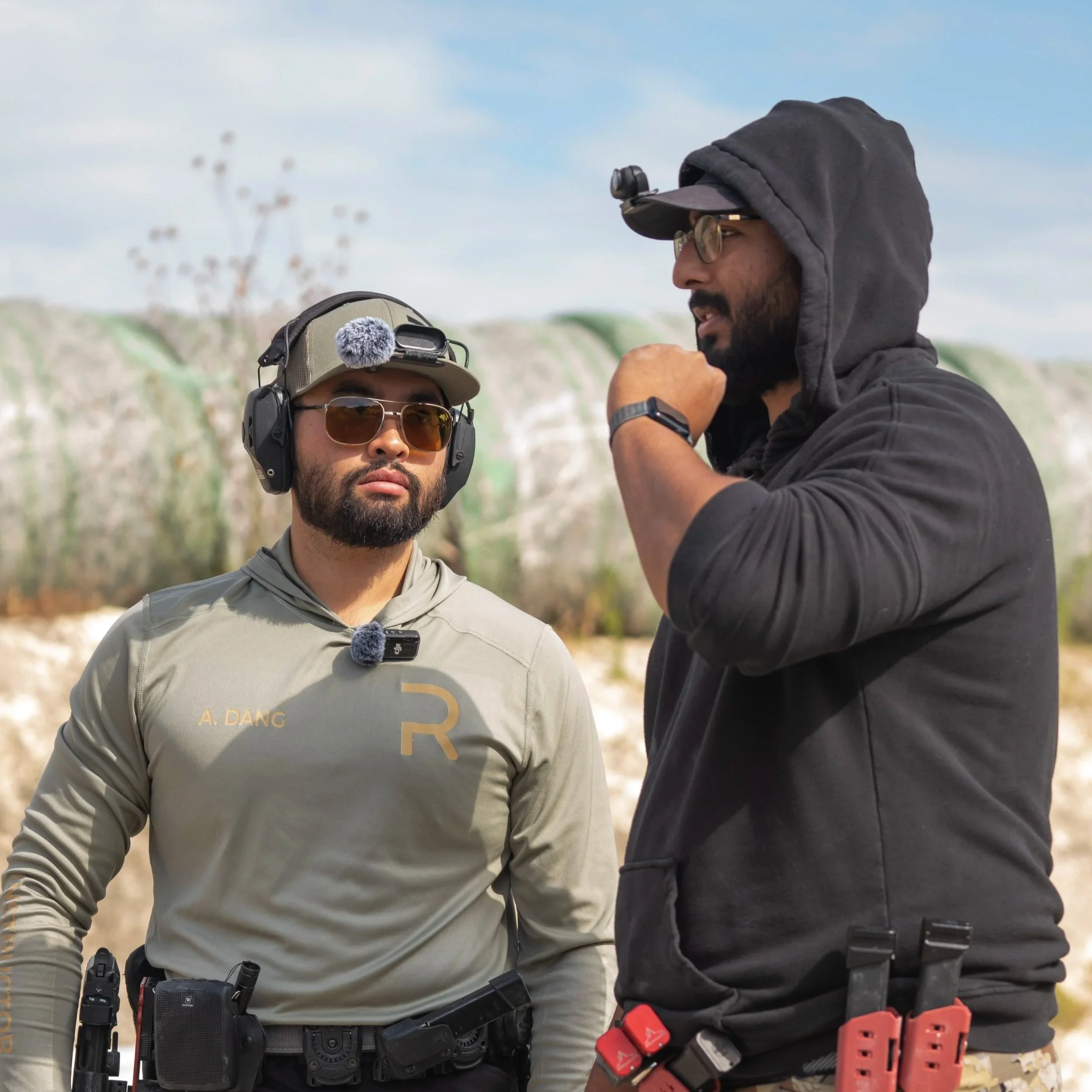 Two men engaging in a discussion outdoors with cliffs in the background. One man is wearing a hat, sunglasses, and a tactical vest with equipment, while the other is dressed in a black hoodie, glasses, and has a headlamp. They appear to be involved in an outdoor activity such as shooting or training.