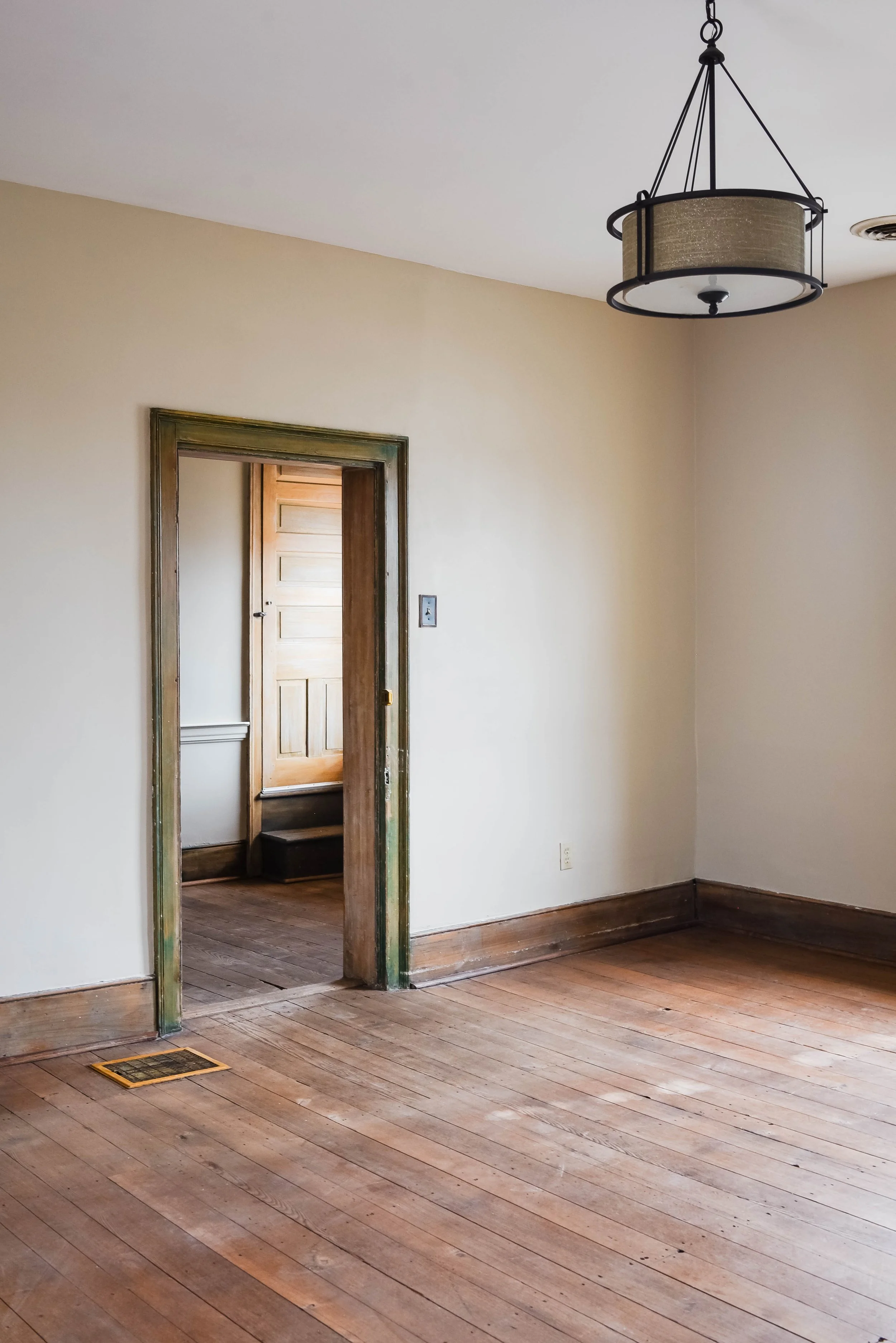 Empty room with wooden floor and open doorway leading to another room, featuring a rustic door and ceiling light fixture.