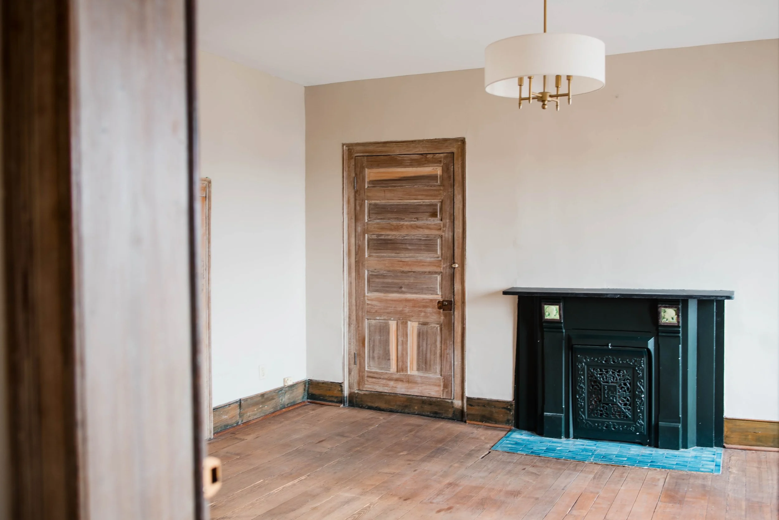 Empty room with wooden floor, light-colored walls, wooden door, decorative black fireplace, and a white ceiling lamp.