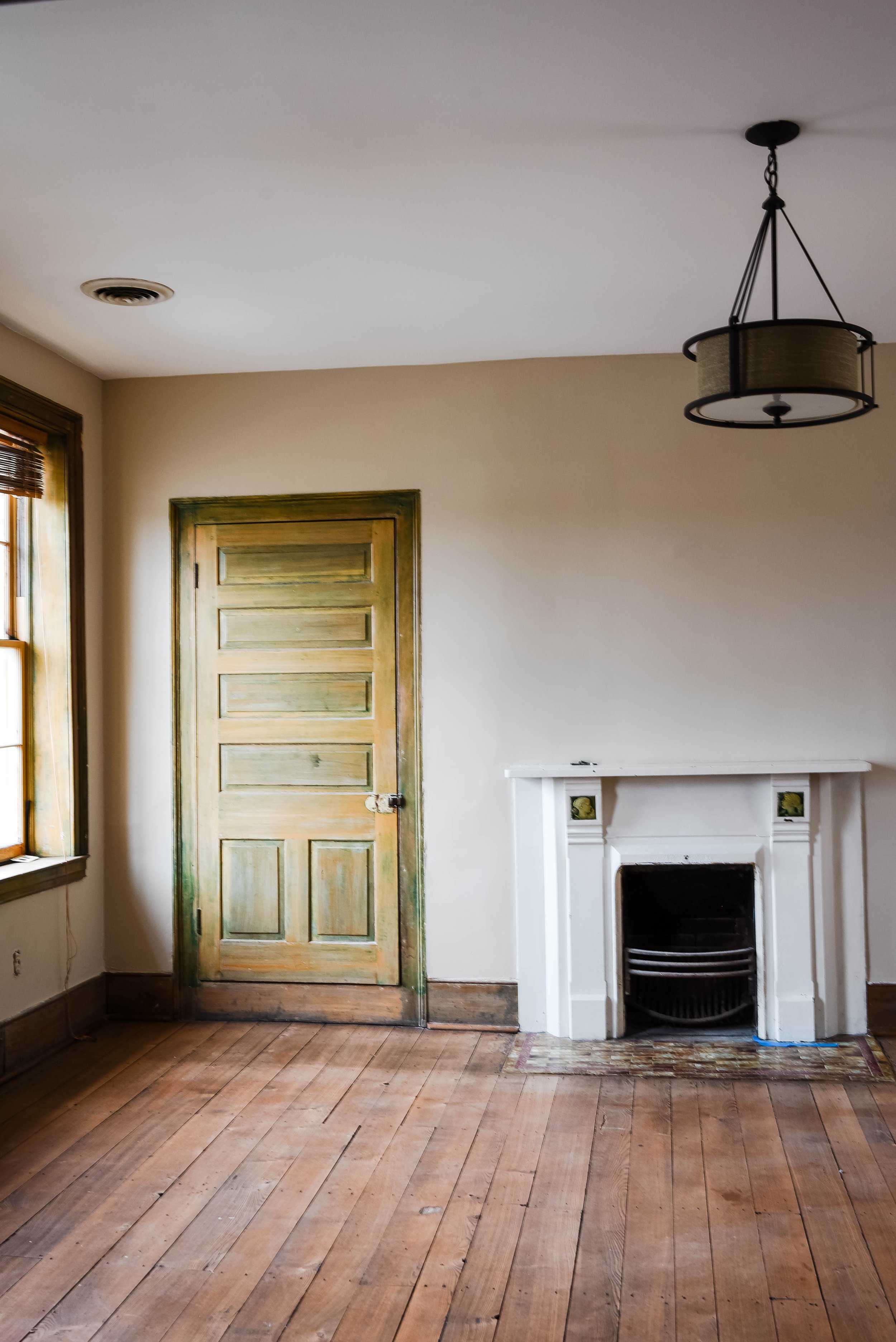 Empty room with wooden floor, a wooden door, fireplace, window, and ceiling light fixture.