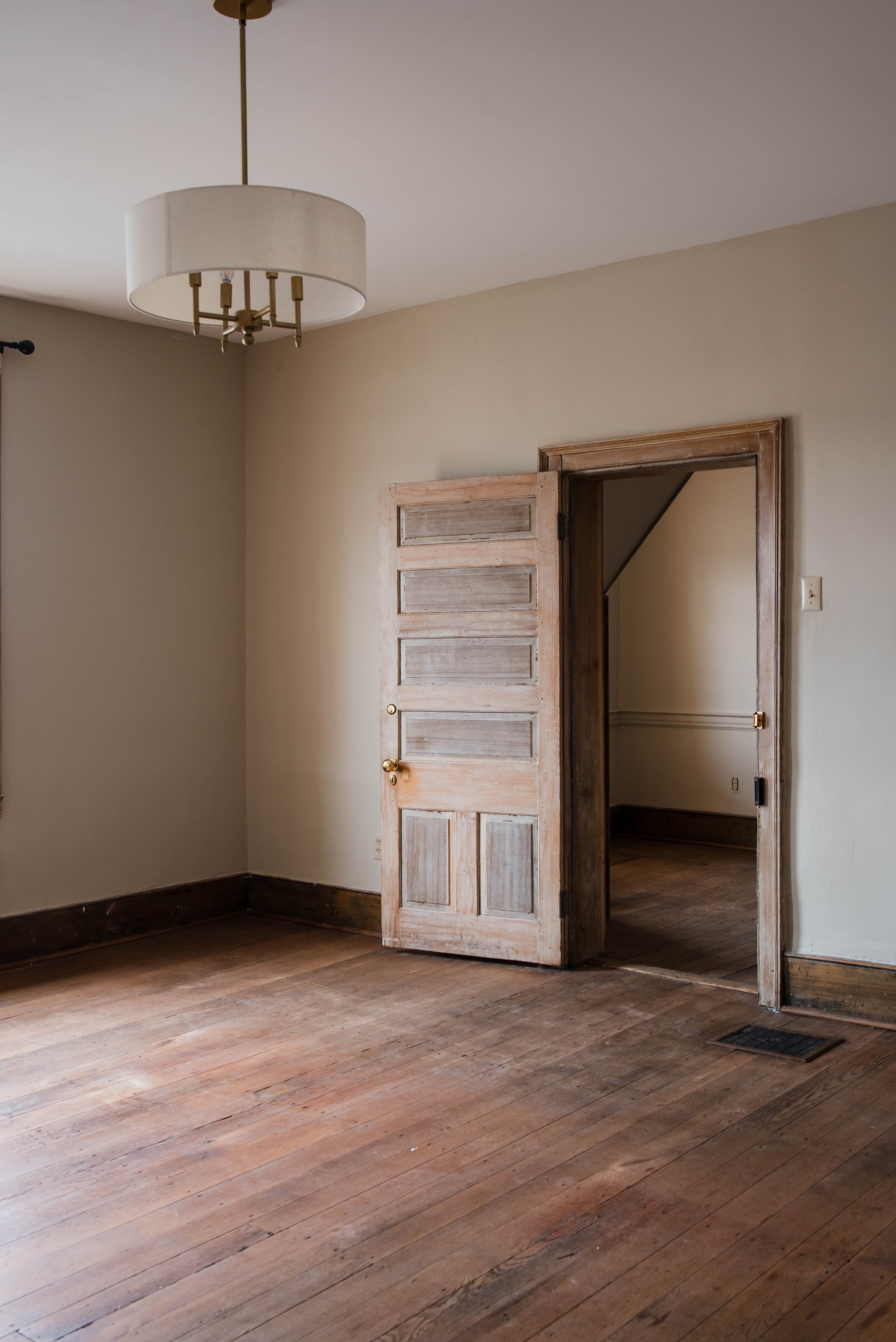 Empty room with wooden floor, beige walls, an open wooden door, and a ceiling light fixture.