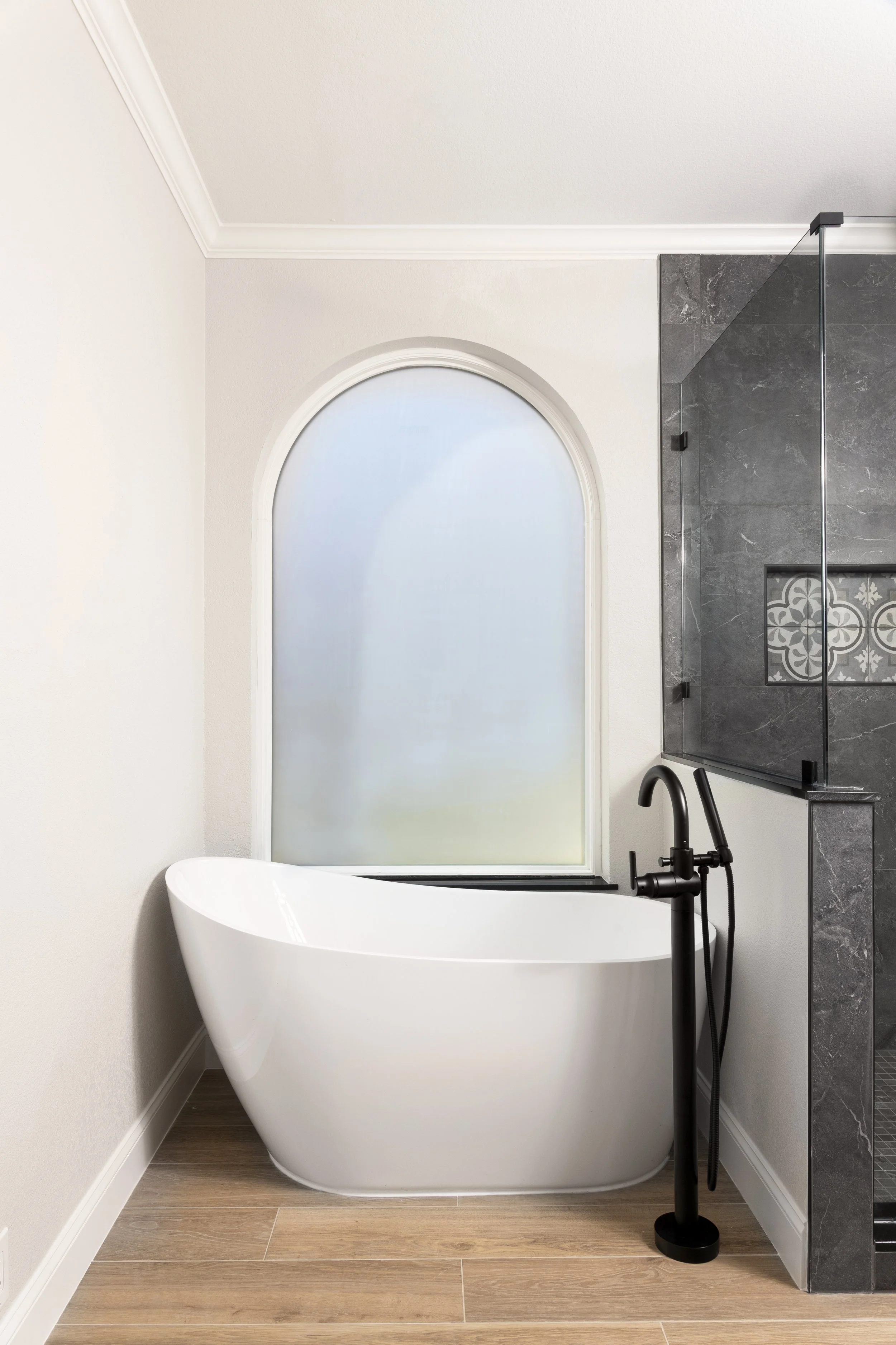 Modern bathroom with white freestanding bathtub, large frosted window, black floor-mounted faucet, and a glass-enclosed shower with dark gray marble tiles.