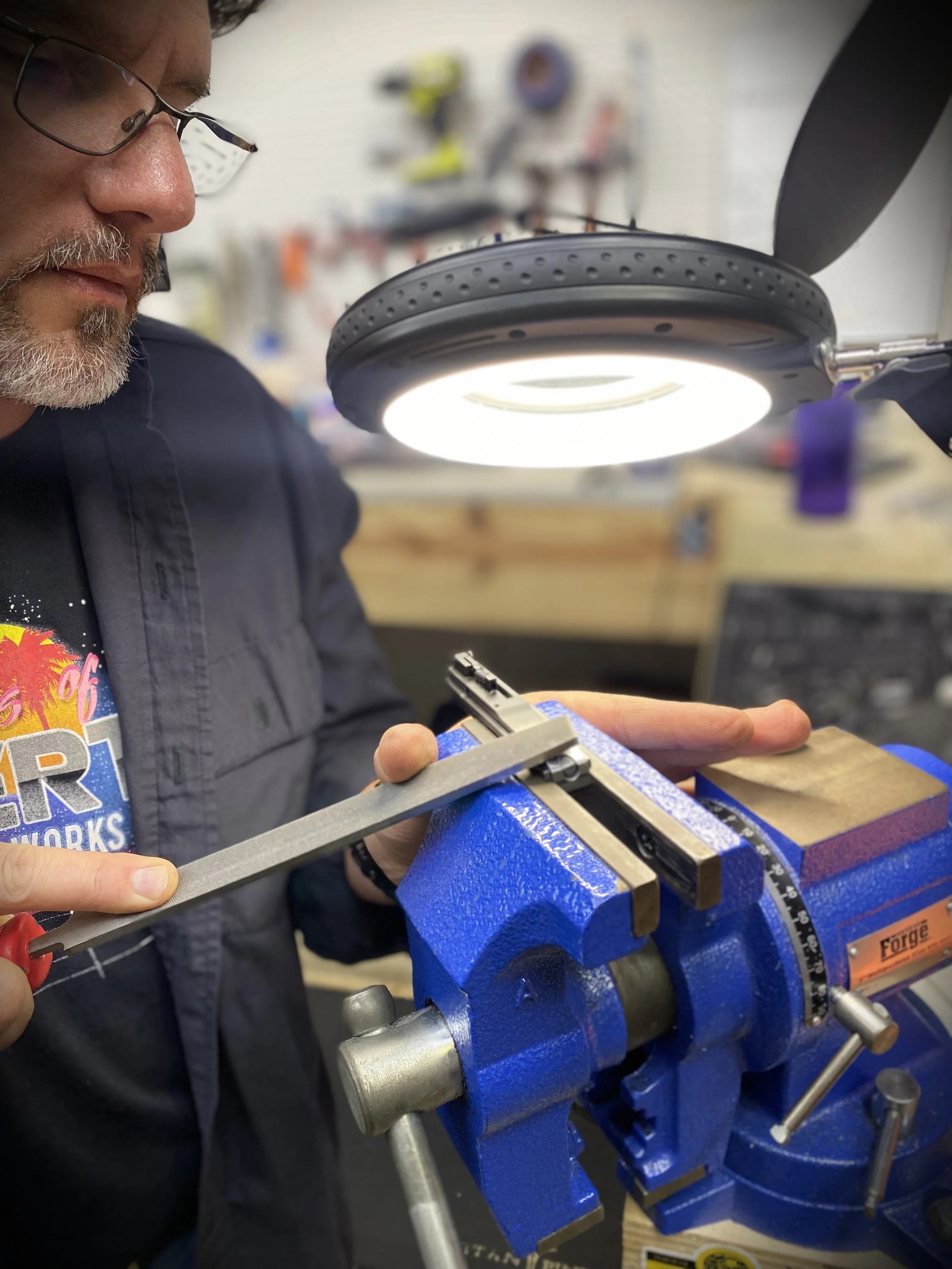 Tex-Star Gunworks in Plano Texas. Cerakote, Gunsmith, and Firearm Services.. Chuck Girard working on a metal project in a workshop, using a blue vice and a magnifying lamp.