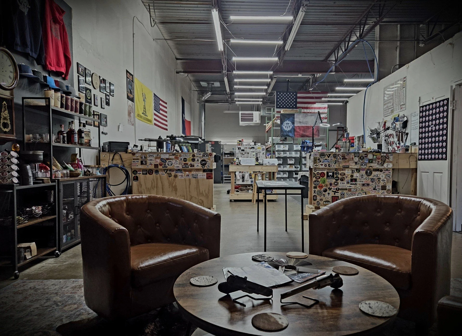 A workshop or office space with two brown leather armchairs around a round wooden table, holding a gun and magazines. In the background, shelves, flags, and various tools and equipment are visible.